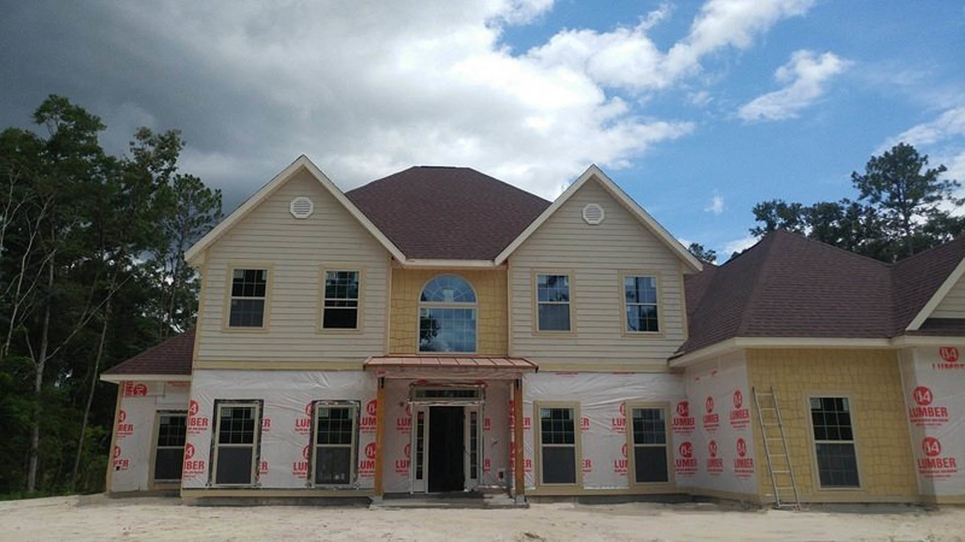 Partially built house with white framed windows, black door with white trim, white vent on exterior wall, red and white construction signs, ladder leaning against wall, trees and