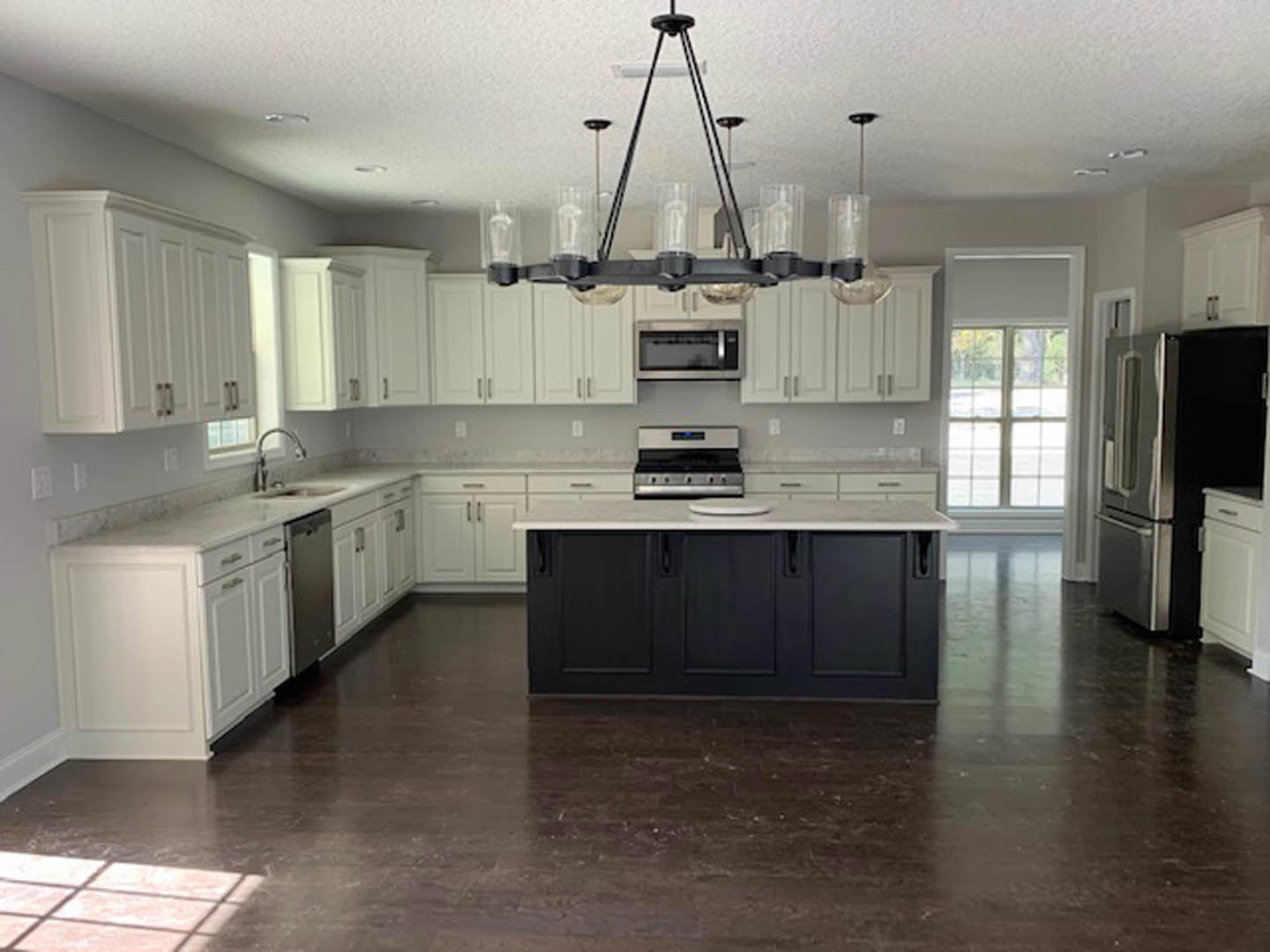 White kitchen with shaker cabinets, black island with quartz countertop, stainless steel refrigerator, built-in microwave, farmhouse sink, light wood flooring, recessed lighting.