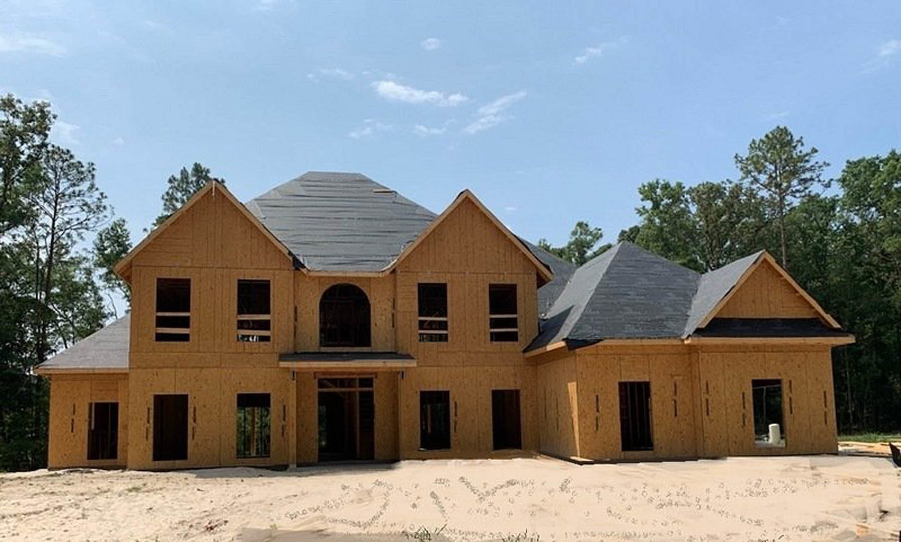 Custom home under construction on sandy ground with large grey octagonal roof, black roofing material, white sheet with writing, dark arched doorway with window, and barred window.