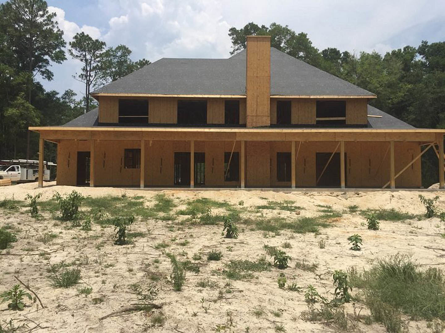 Framed house under construction with exposed wood beams, chimney, and unfinished roof, surrounded by dirt field with scattered plants under cloudy sky