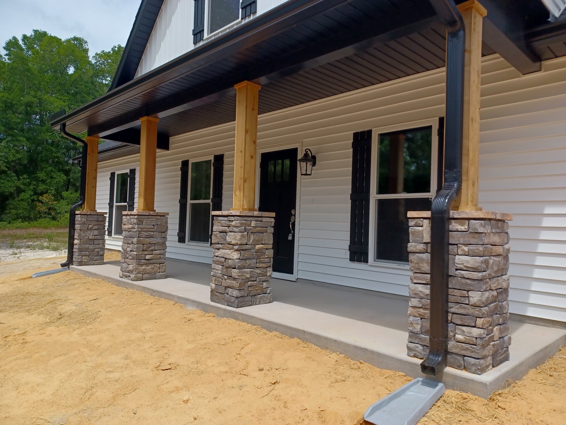 Stone pillars frame the porch of a custom home with black and white siding, a dirt path leading to the entrance, stacked bricks and a plastic container near the door, and trees