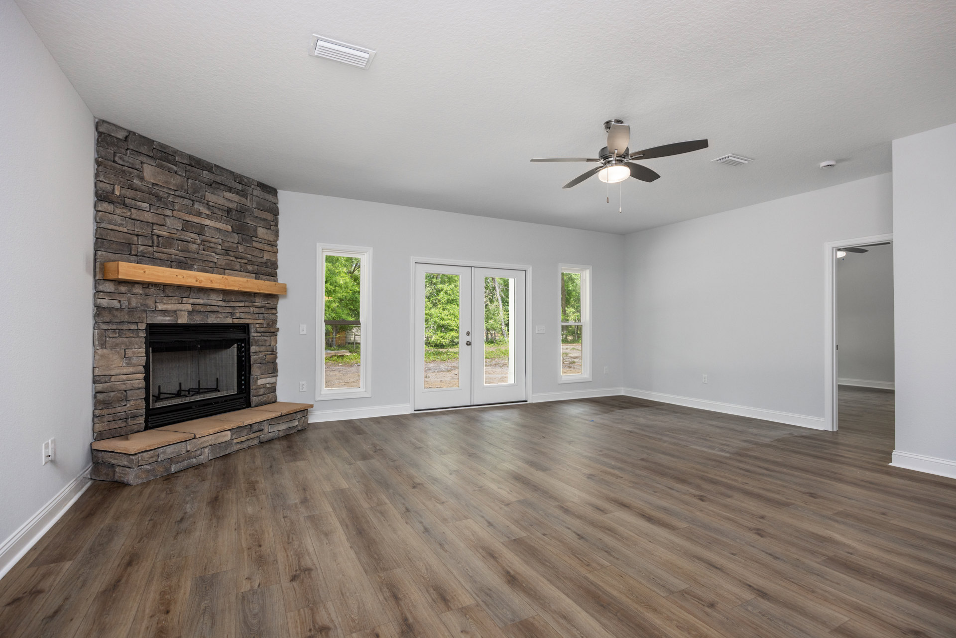 Living room with wood flooring, stone fireplace wall, wood mantel shelf, ceiling fan with light, double glass-paneled doors, and plaster walls.