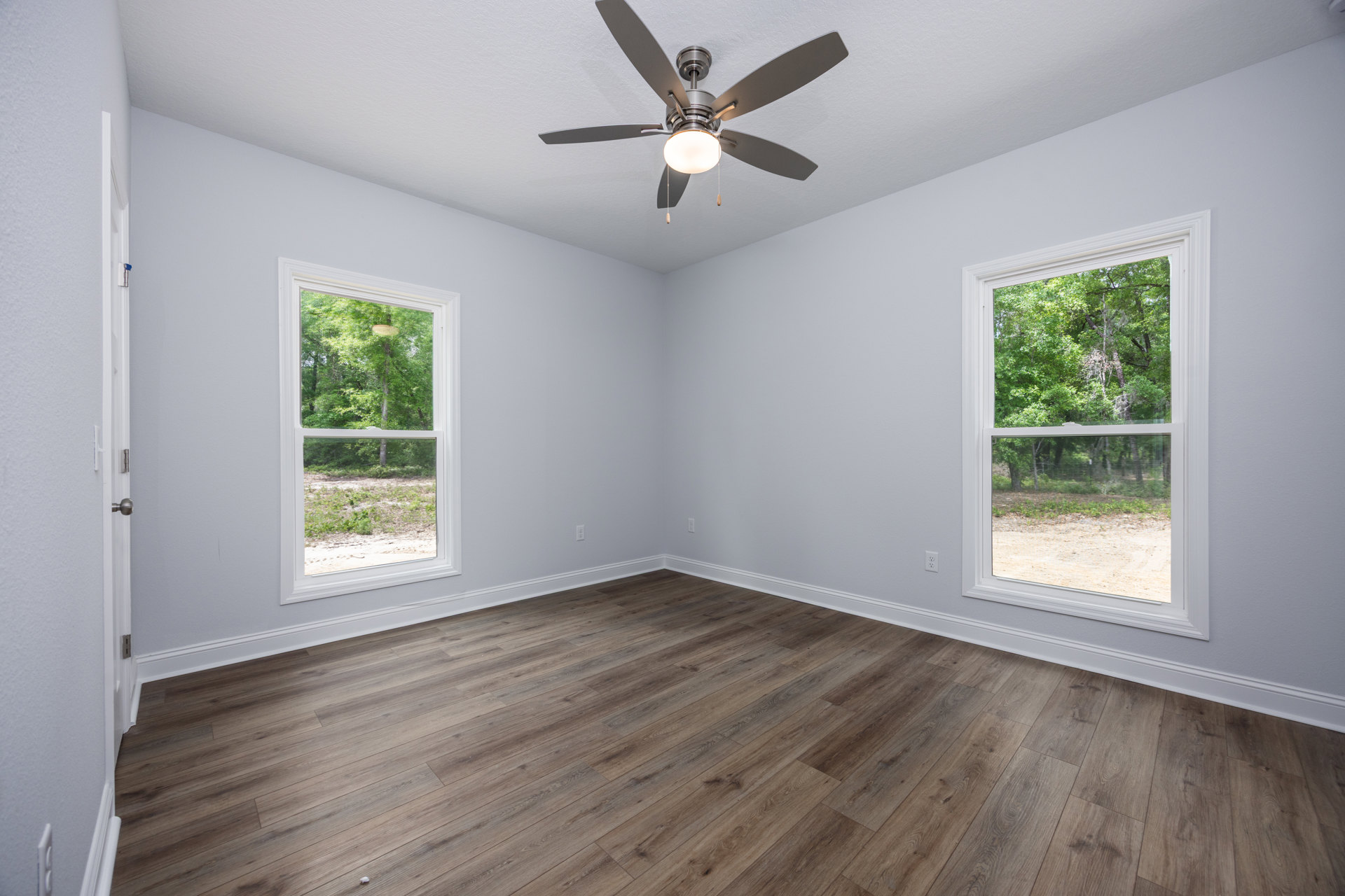 Ceiling fan with light fixture above wood flooring, large windows with tree views, white plaster walls and ceiling