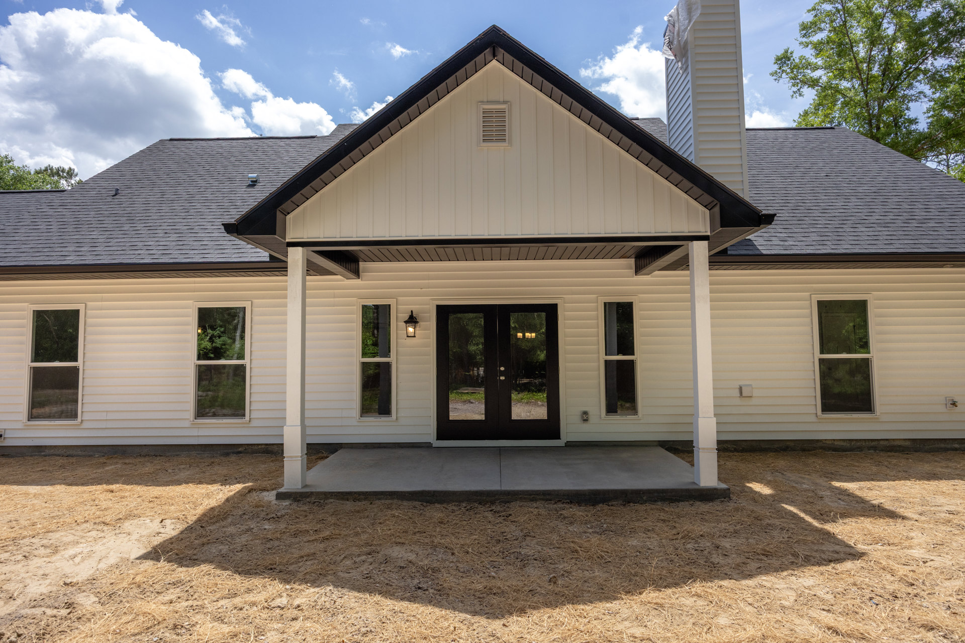 White siding house with black shingle roof, double glass-paneled doors, white-framed windows, covered porch, large white tower feature, vent detail, and hay-covered ground.