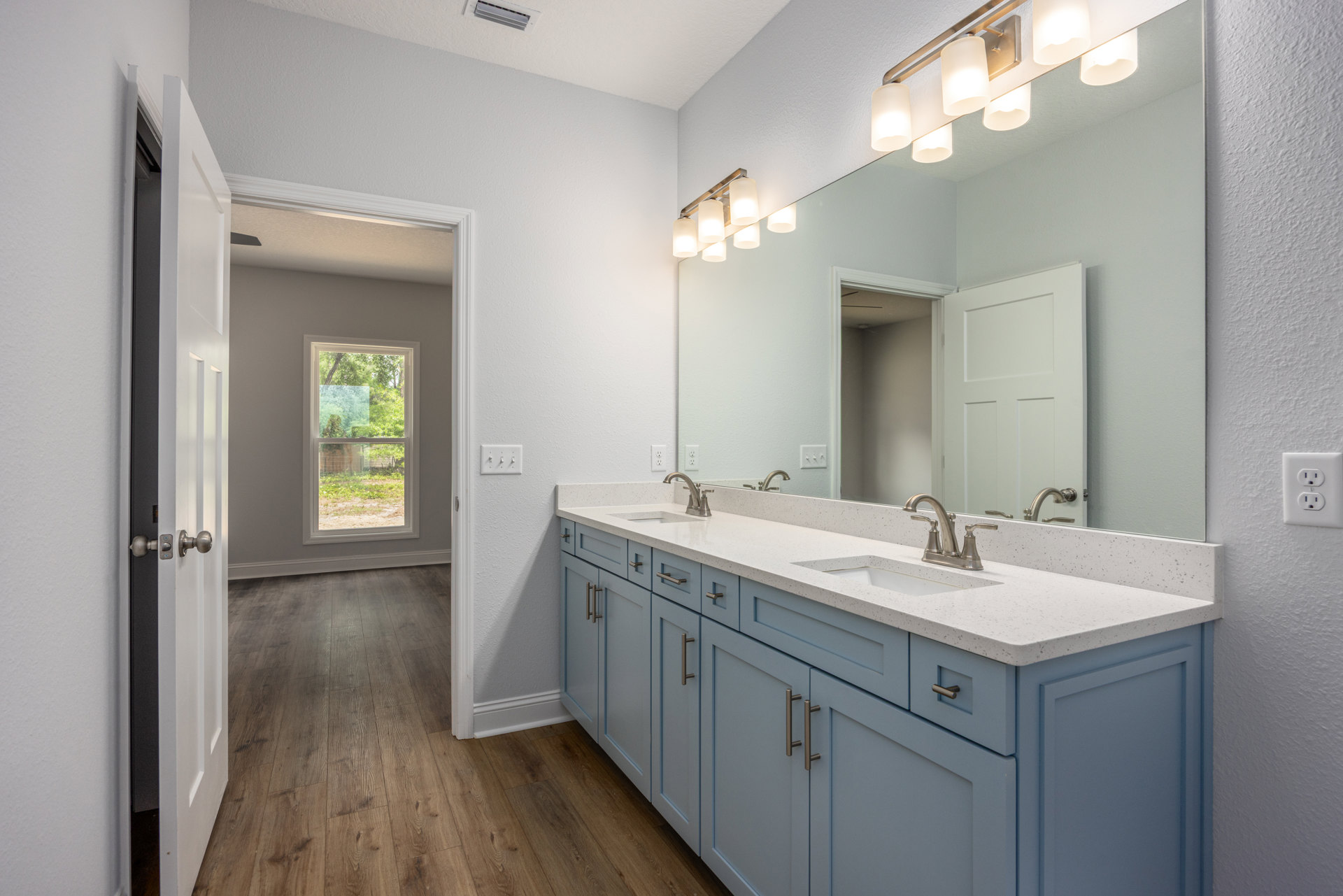 Bathroom with blue shaker cabinets, wide vanity mirror, white countertop, chrome faucet, wall-mounted light fixture, tile flooring, and window overlooking trees.