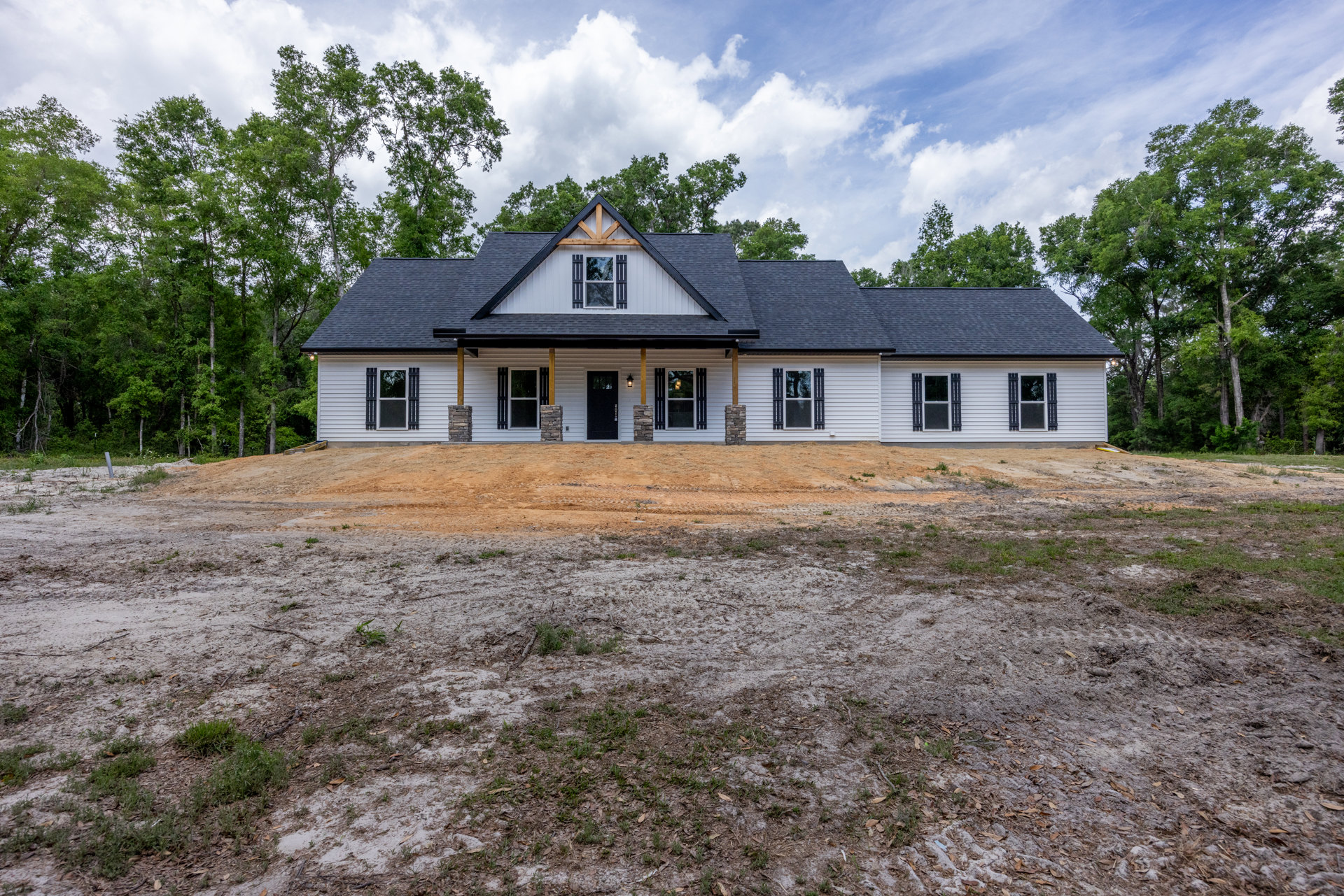 Two-story farmhouse under construction with stone pillars, black front door, white-framed windows, dirt yard, and surrounding trees