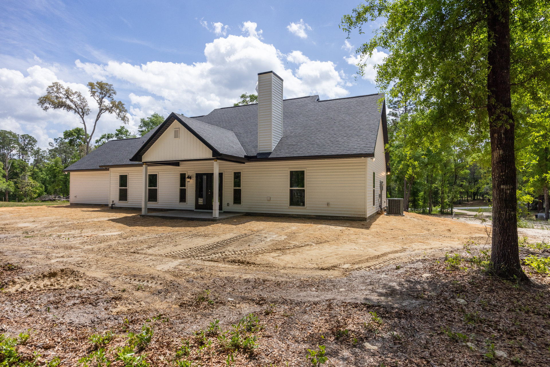 White chimney on gray roof, white-framed window, large metal utility box on concrete base, dirt yard in front of house, cloudy sky and trees in background