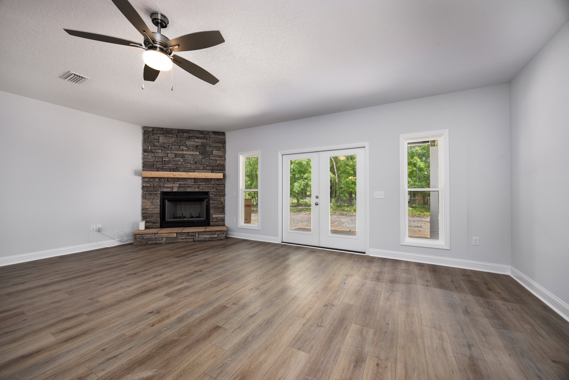 Living room with stone fireplace, wood flooring, ceiling fan with light, white double doors with glass panes, window showing trees outside