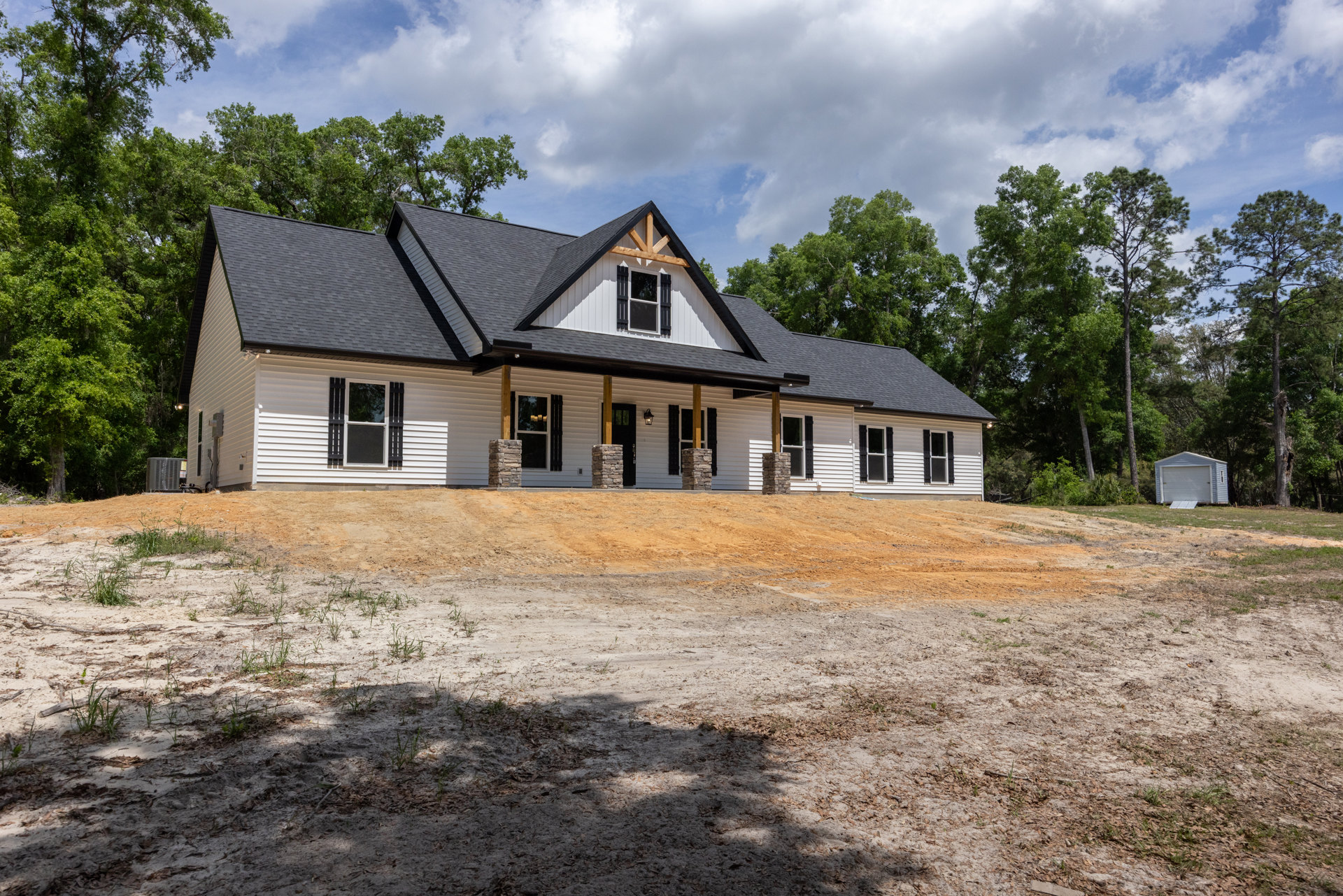 White siding house with black roof, black shuttered windows, dirt driveway and dirt hill in front, small shed with door to the side, rolls of toilet paper visible in window