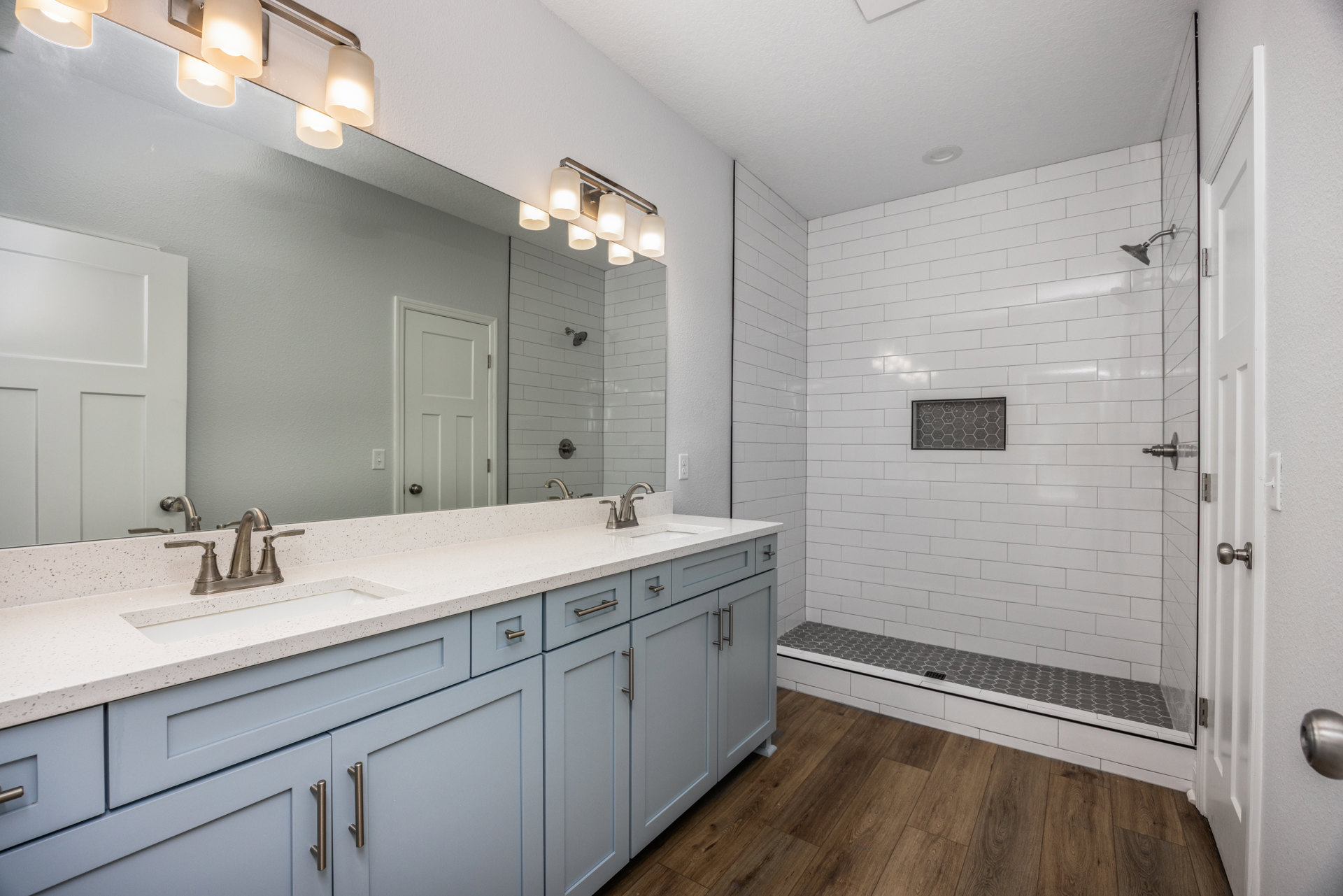 Bathroom featuring a large wall mirror, glass-enclosed shower, white door with metal frame, light fixture with multiple bulbs, tiled walls and floor, metal hardware, and