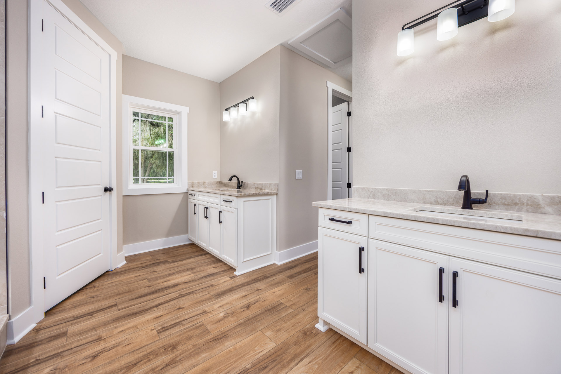 Bathroom with white cabinets, wood flooring, marble countertop with black faucet, window showing trees, white lamp shade on black surface, white door with black knobs and hinge