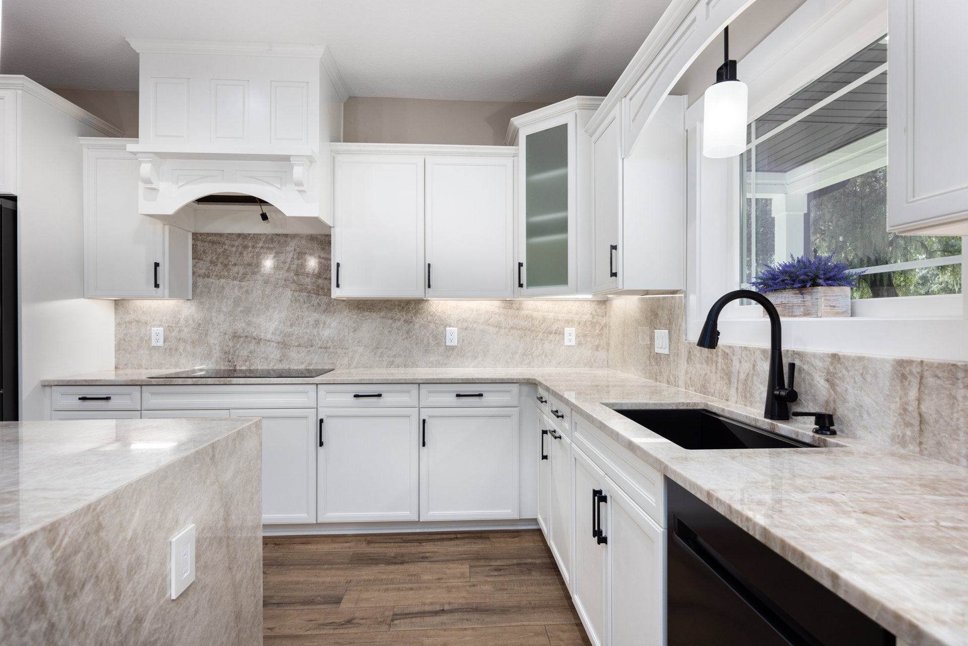 White kitchen cabinets and marble countertops, black sink with matching faucet, planter of purple flowers on the counter, white outlet set into marble, white door with black handle