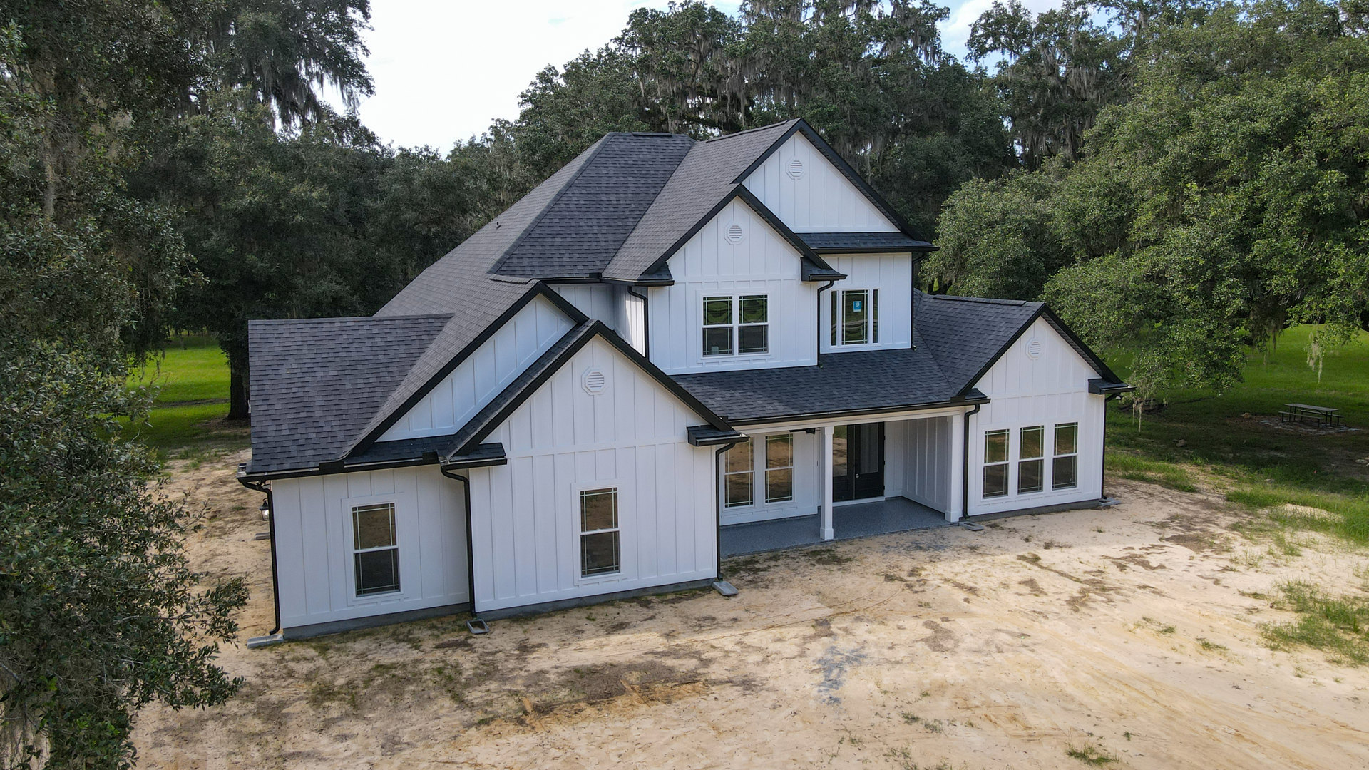 White two-story house with multiple windows, surrounded by mature trees and green foliage, dirt ground in front, blue sign visible on glass door