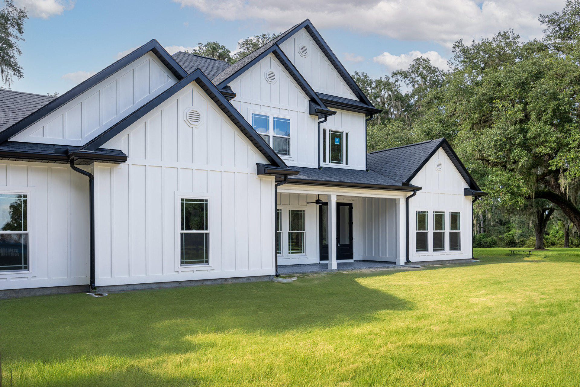 White house with black roof, white-framed windows reflecting trees, white wall vent, green lawn in front, partly cloudy sky overhead.