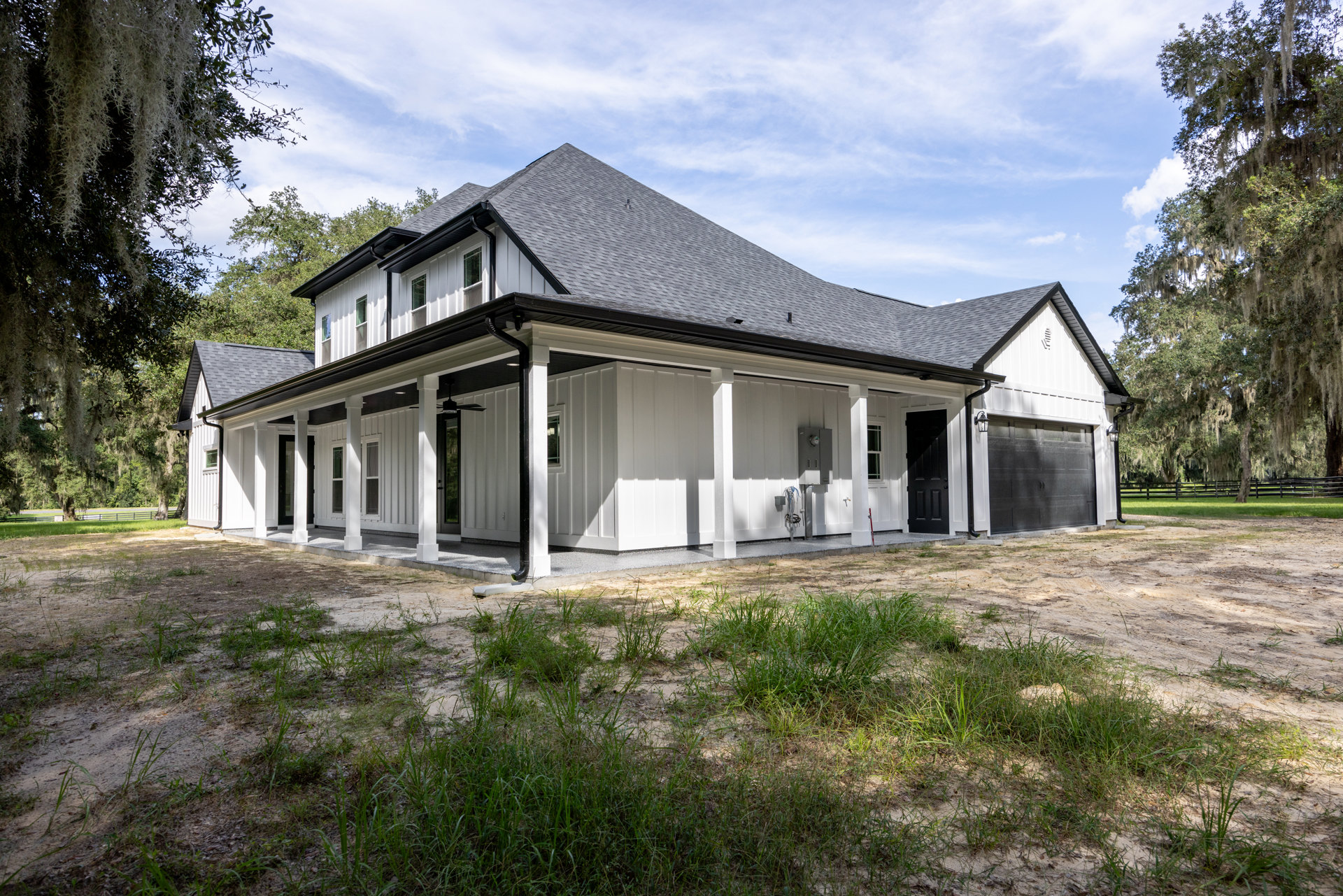 White two-story house with attached garage, gray shingle roof, covered front porch, manicured lawn, and mature tree with moss in foreground.