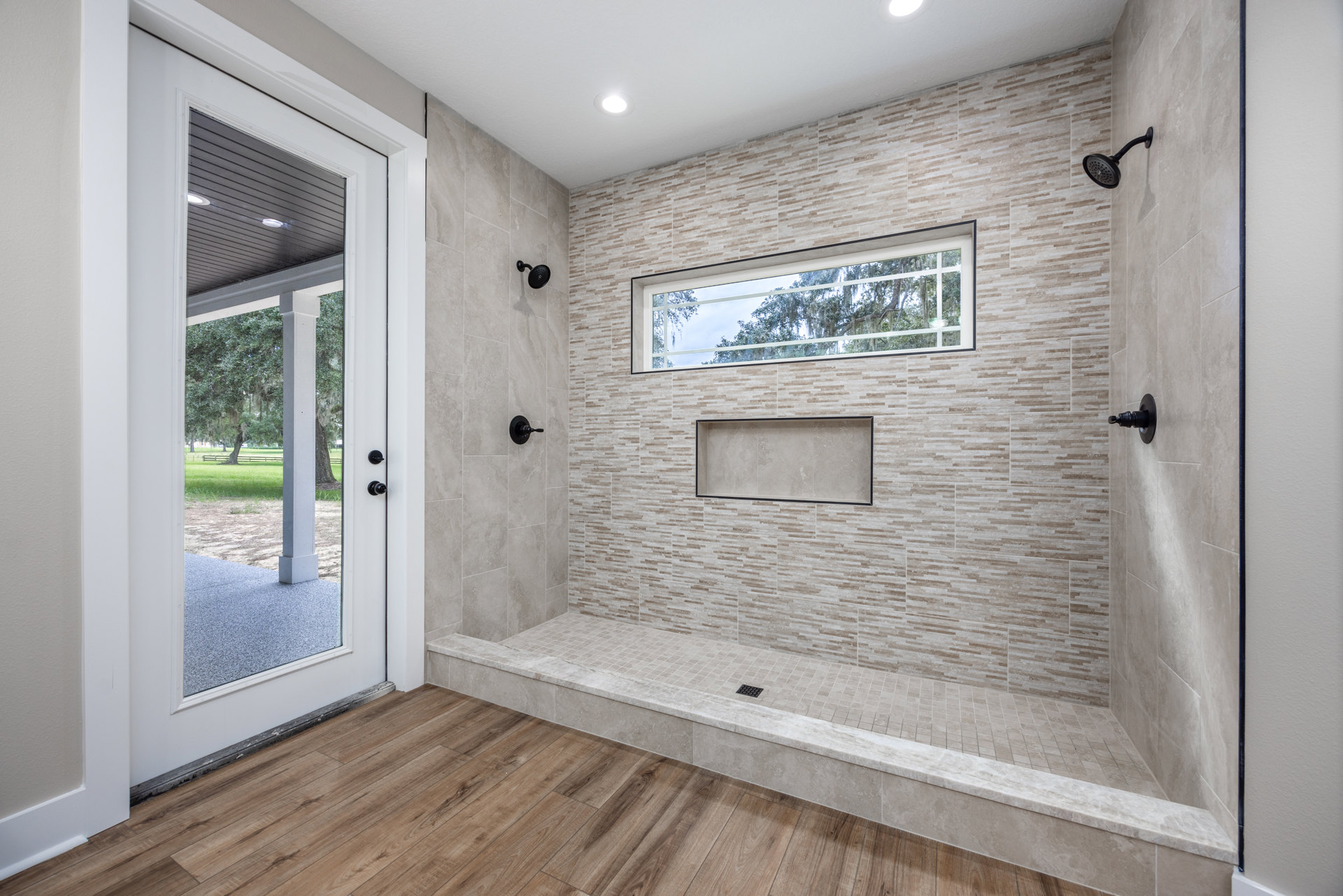 Bathroom with rectangular white tile and black trim, glass shower door, black shower head and pole, window overlooking trees, door with view of grass field and trees, laminate