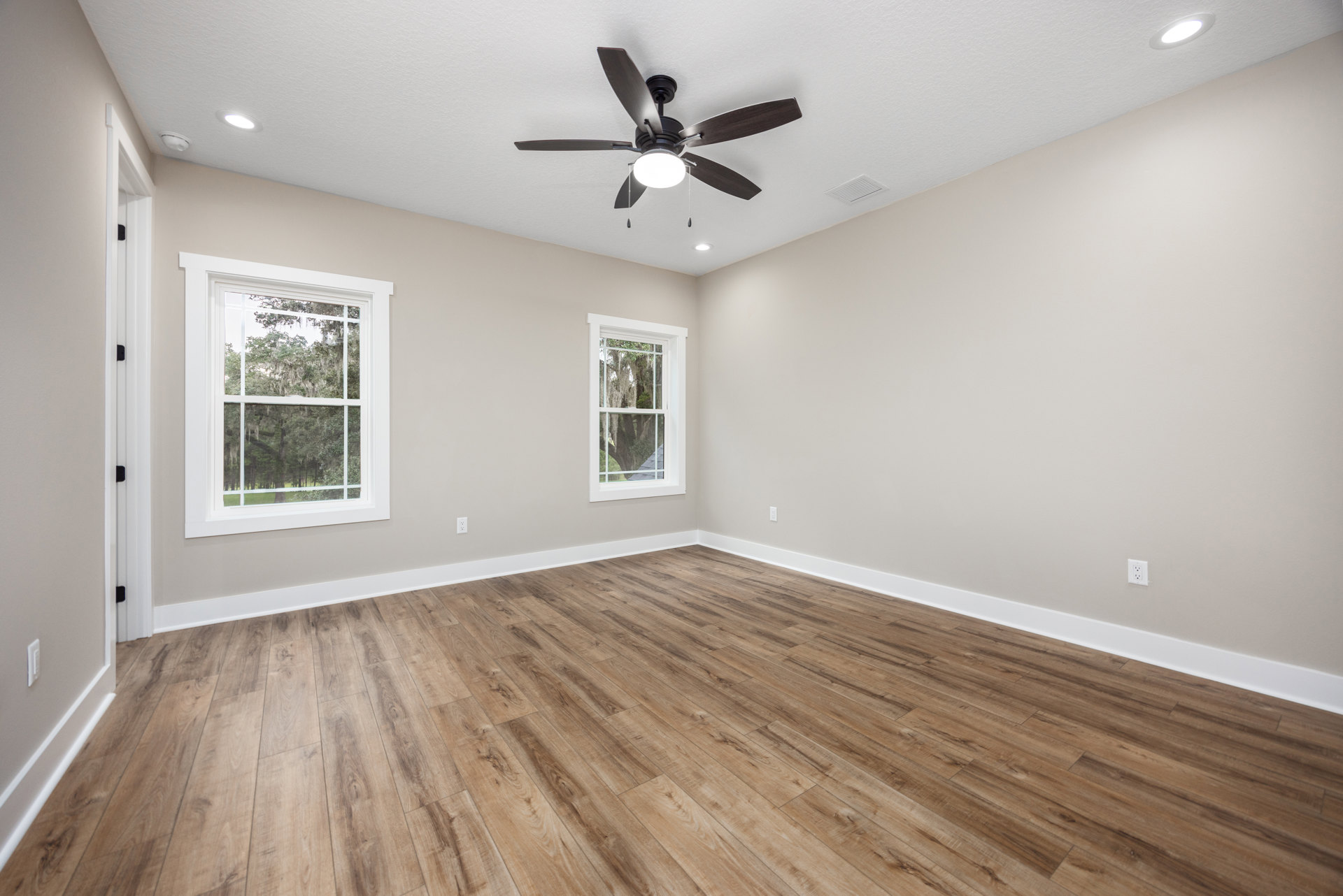 Ceiling fan with light fixture above wood flooring, white trim, and two windows with white frames overlooking trees