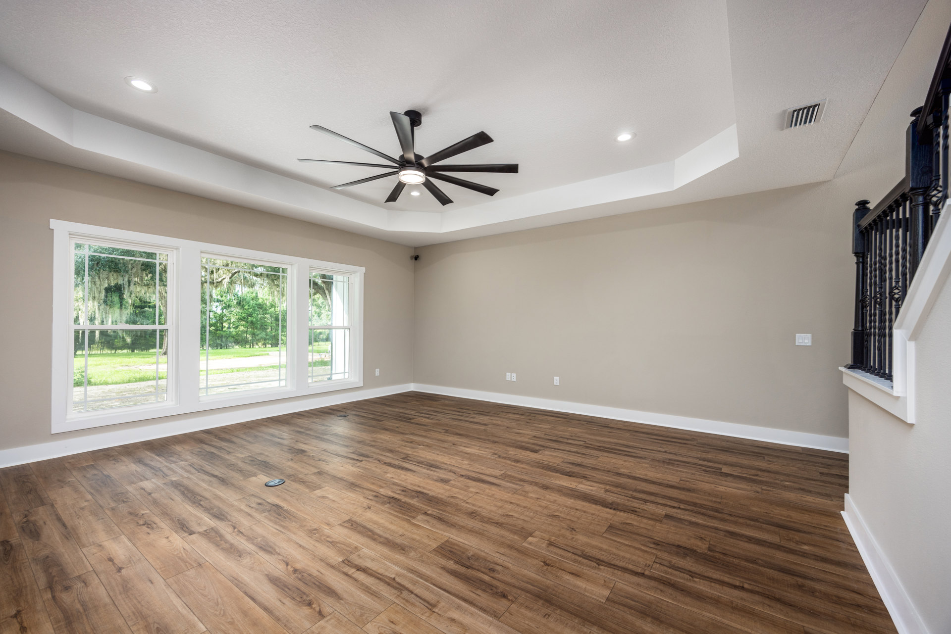 Living room with wood flooring, white walls, ceiling fan with light, large windows overlooking trees and grass, black metal railing in foreground