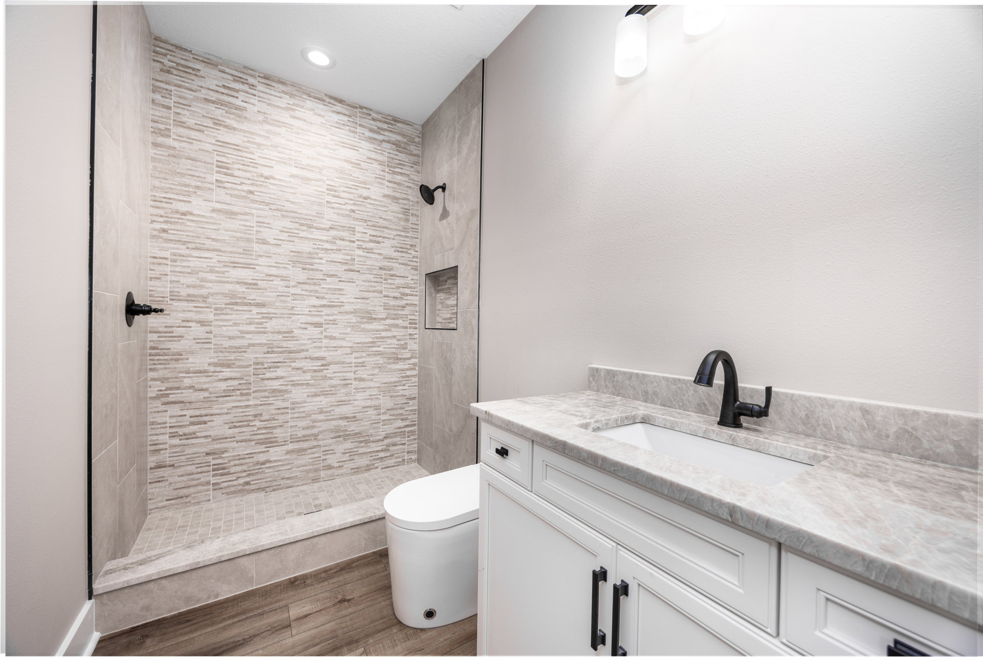 Bathroom with marble countertop and black faucet, white toilet, glass-enclosed shower with tile walls, stone accent wall, and modern white light fixture.