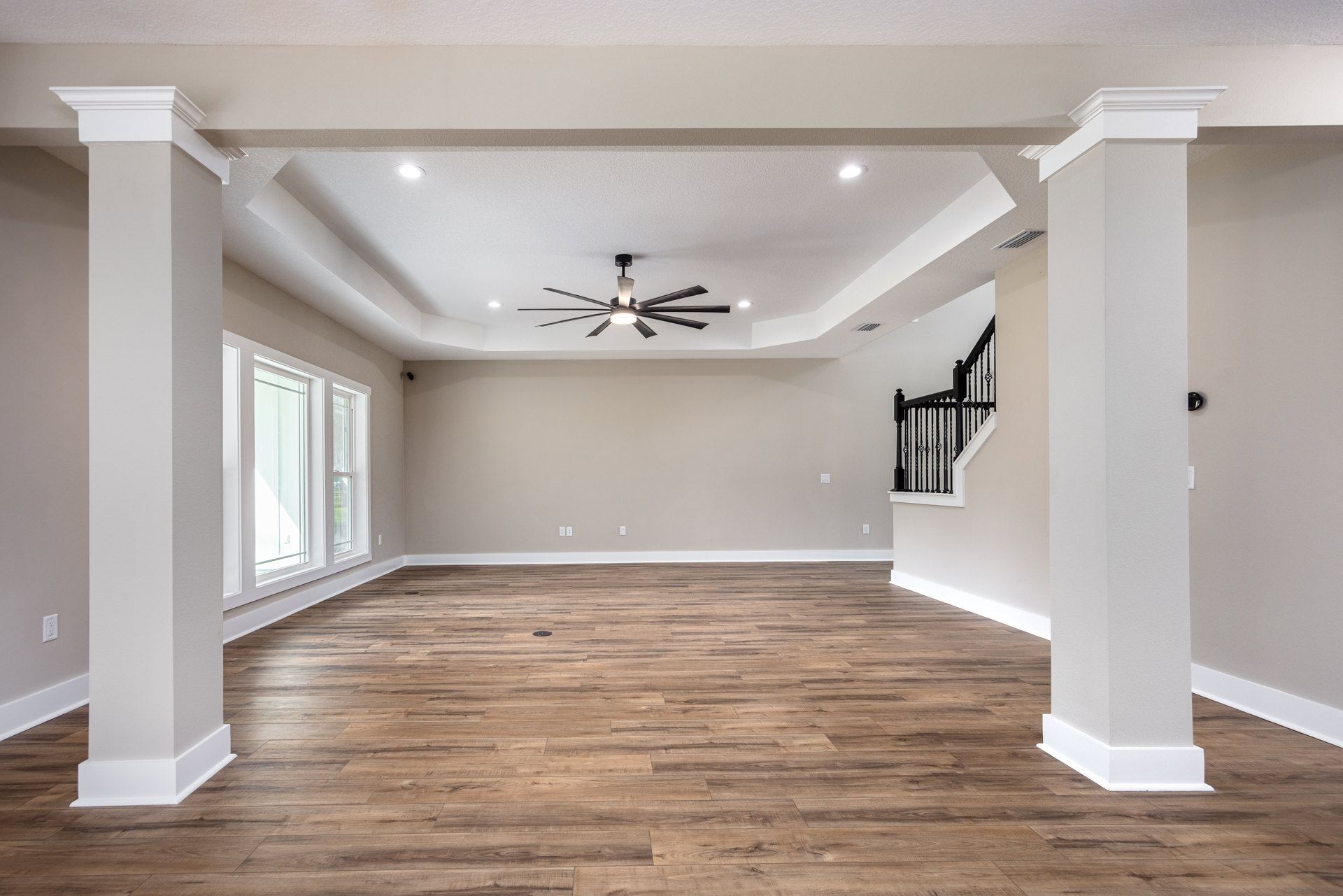 Spacious room with wood flooring, white walls, ceiling fan with light, black staircase railing, and white-framed window