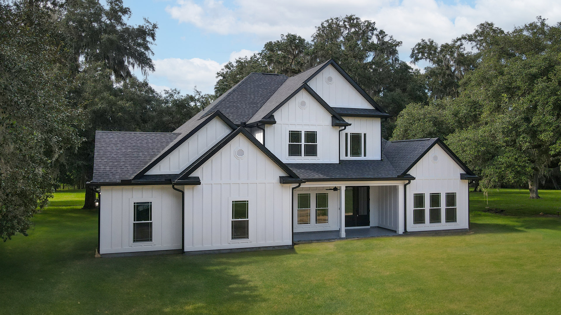 White two-story house with black roof, expansive green lawn, large windows, and mature trees under partly cloudy sky