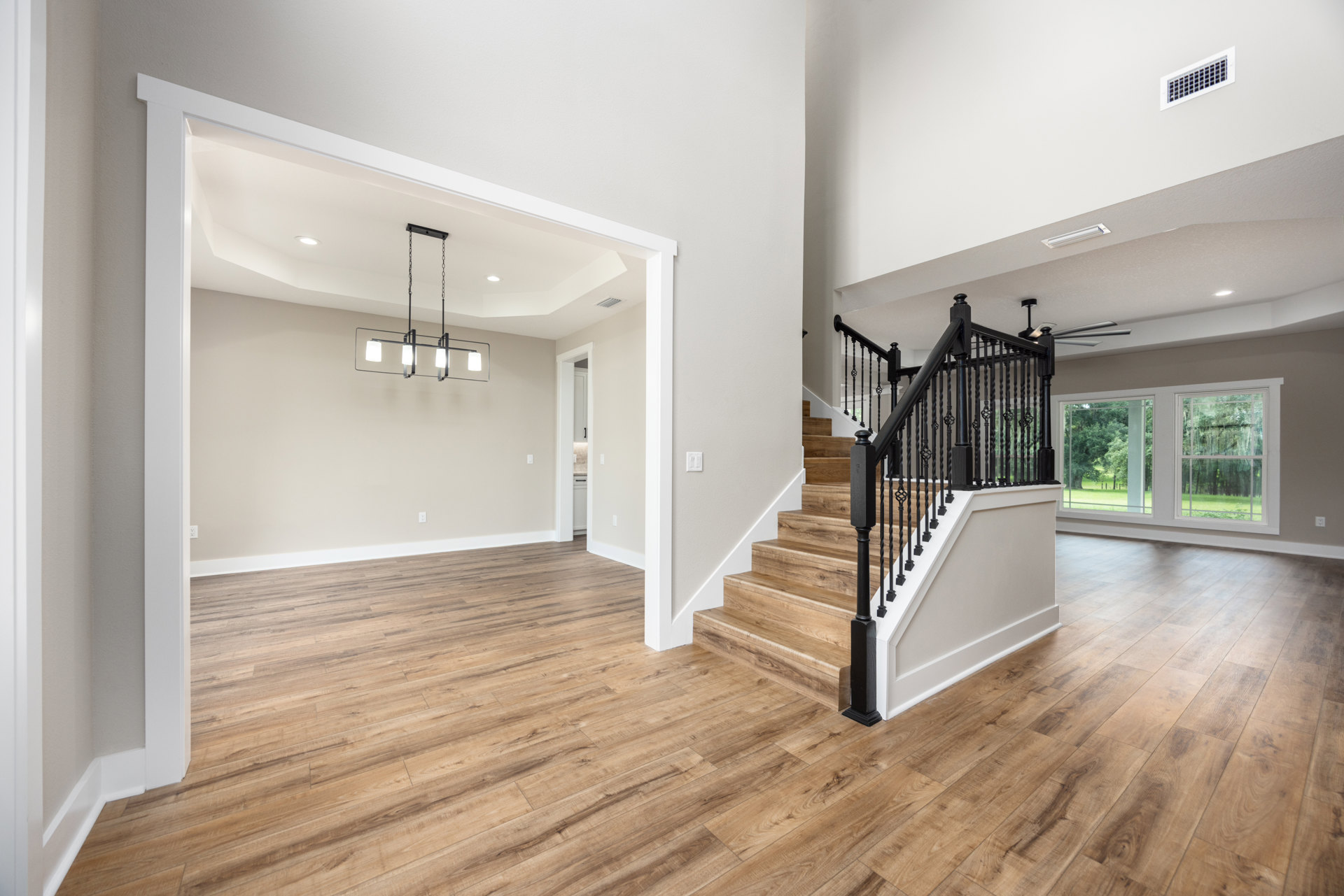 Wood staircase with black metal railings, hardwood floors with white baseboards, modern ceiling light fixture, large window overlooking forest, white air vent, square windows on