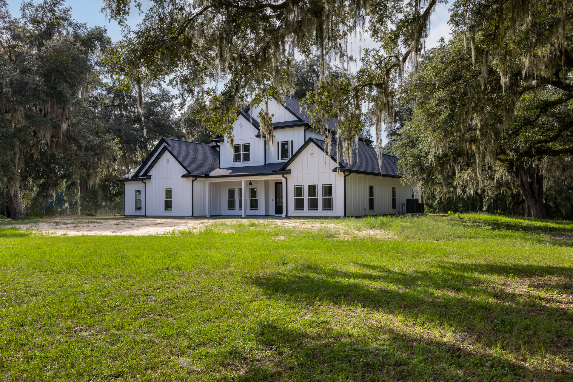 White farmhouse with black-framed windows, surrounded by mature trees, set on a grassy lawn under a clear sky