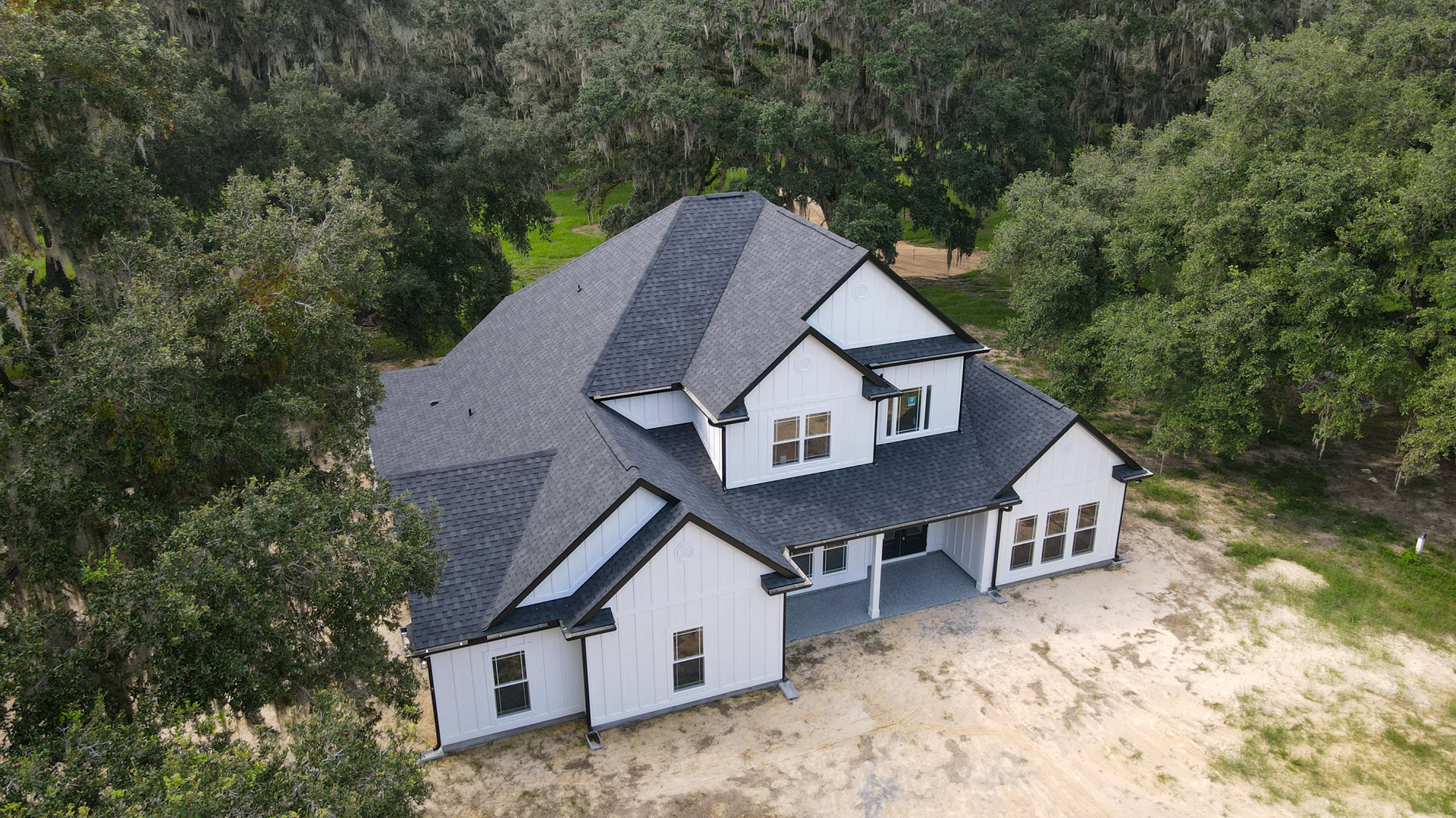 Spacious white home with black roof, expansive driveway, large windows, and mature trees in the background