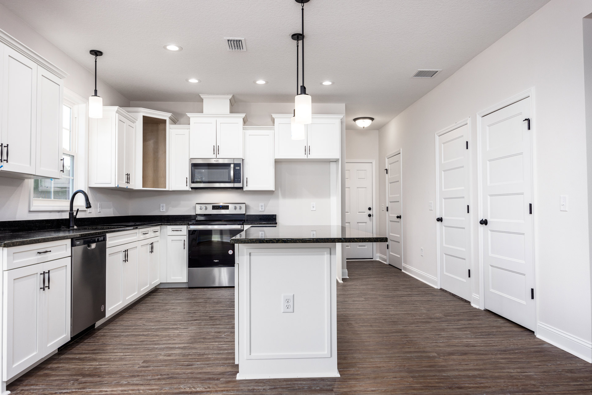 White kitchen cabinets with black marble countertops, black stove and microwave, white door with black knob, power outlet on cabinet, light-colored flooring.