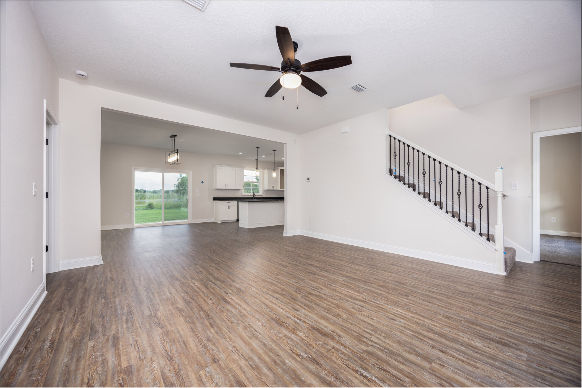 Open living area featuring patterned wood flooring, modern ceiling fan with light, metal-railed staircase, and sliding glass door overlooking grassy field.