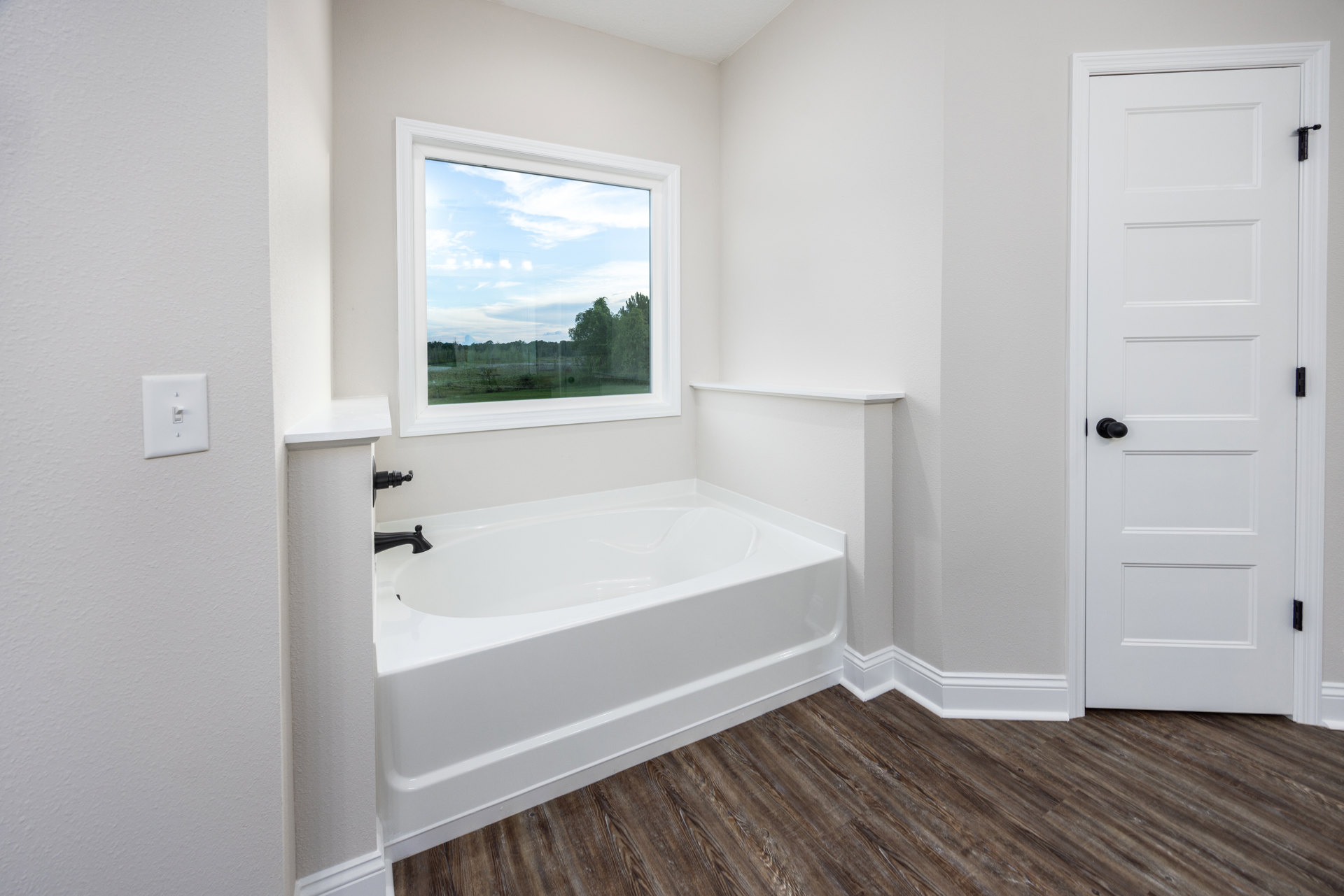 Freestanding white bathtub with black faucet on wood floor, white trim, nearby white door with black knob, window overlooking field and trees, light switch on wall