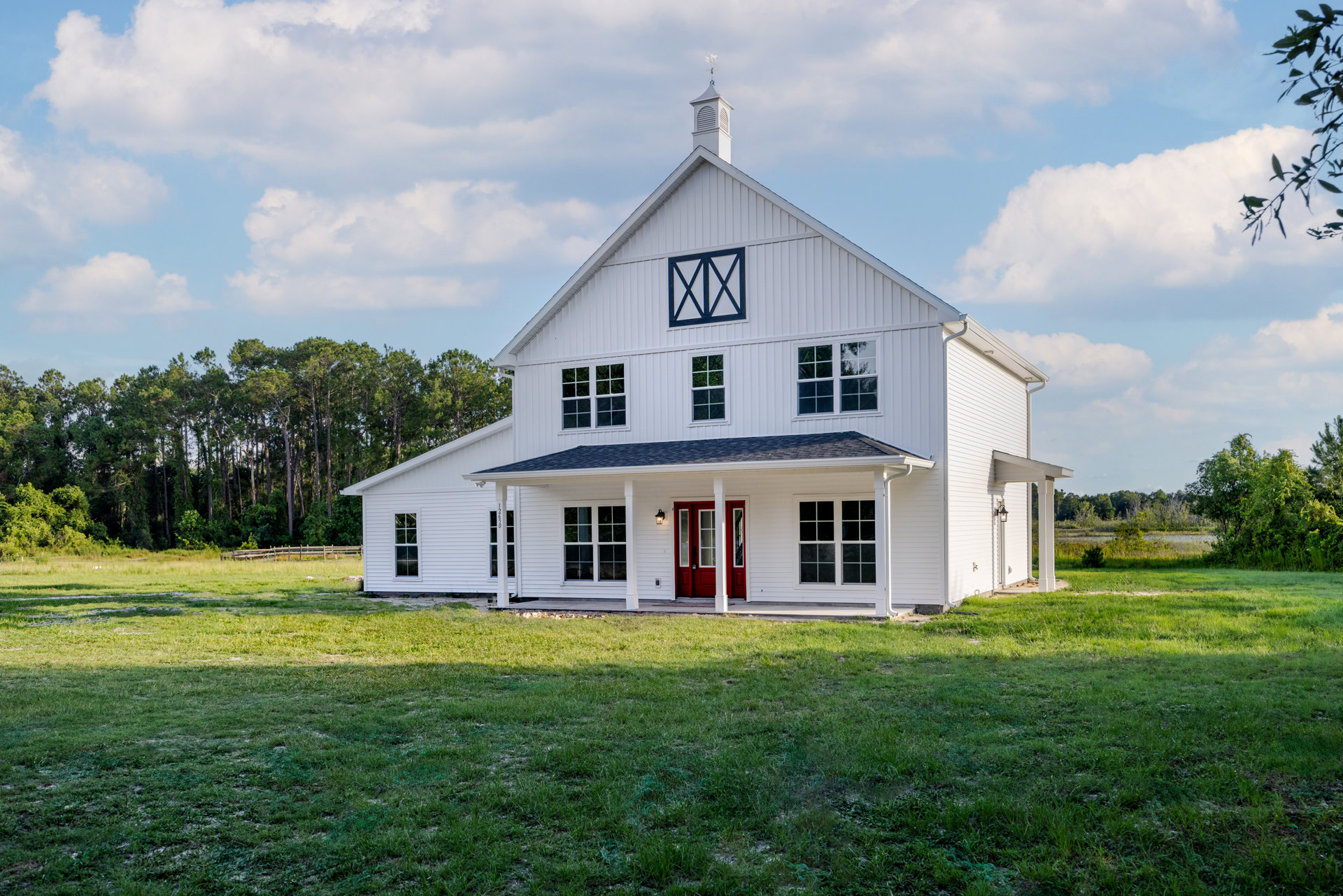 White farmhouse with red front door, white siding, grassy lawn, and trees under a partly cloudy sky