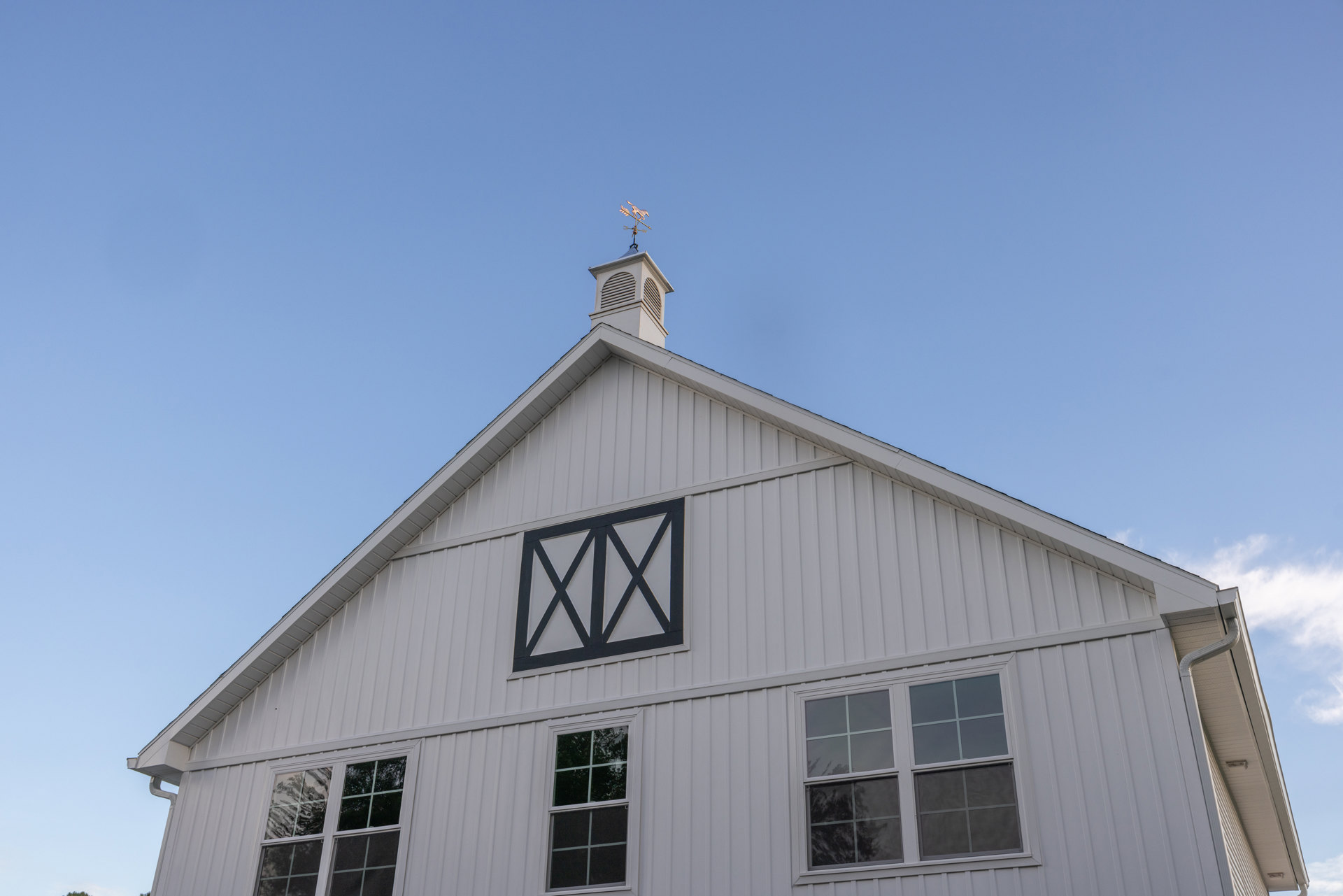 White barn-style home with black square window, white trim, and rooftop weather vane against a partly cloudy sky