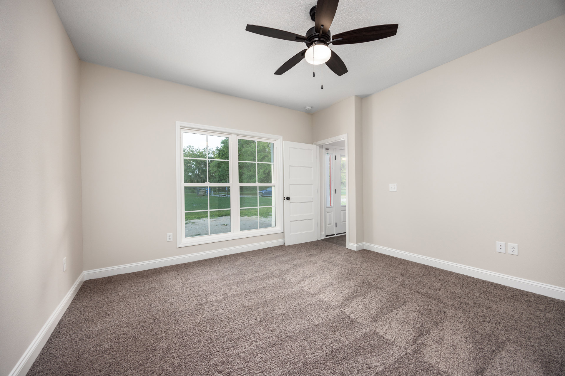Carpeted room with white walls, ceiling fan with light fixture, large window overlooking green yard and trees