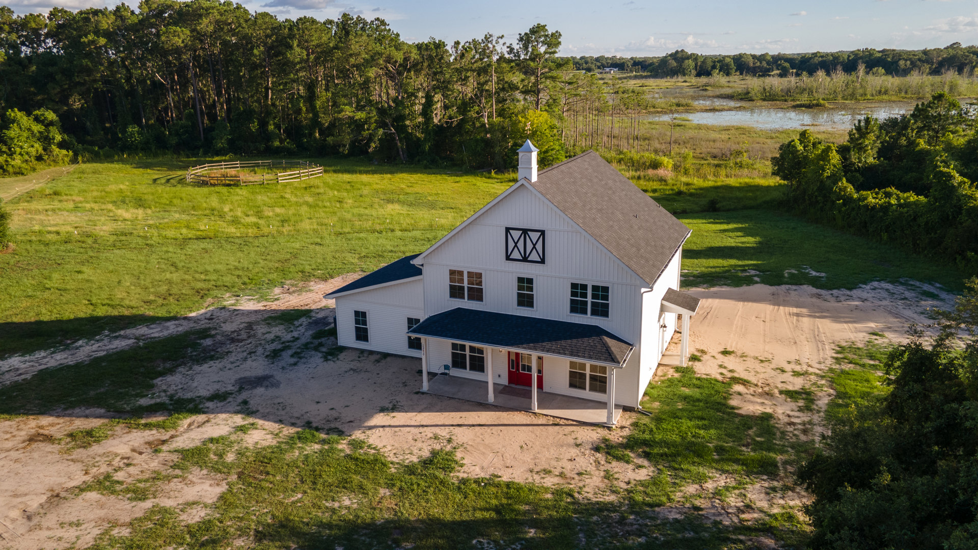 White farmhouse with red door, surrounded by green lawn and large grassy yard, bordered by trees under blue sky