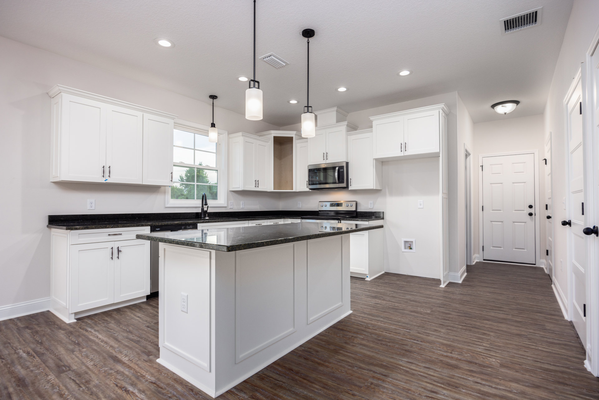 Kitchen with white shaker cabinets, matte black countertops, stainless steel microwave, white door with black handles, kitchen island with black surface, light fixture, and