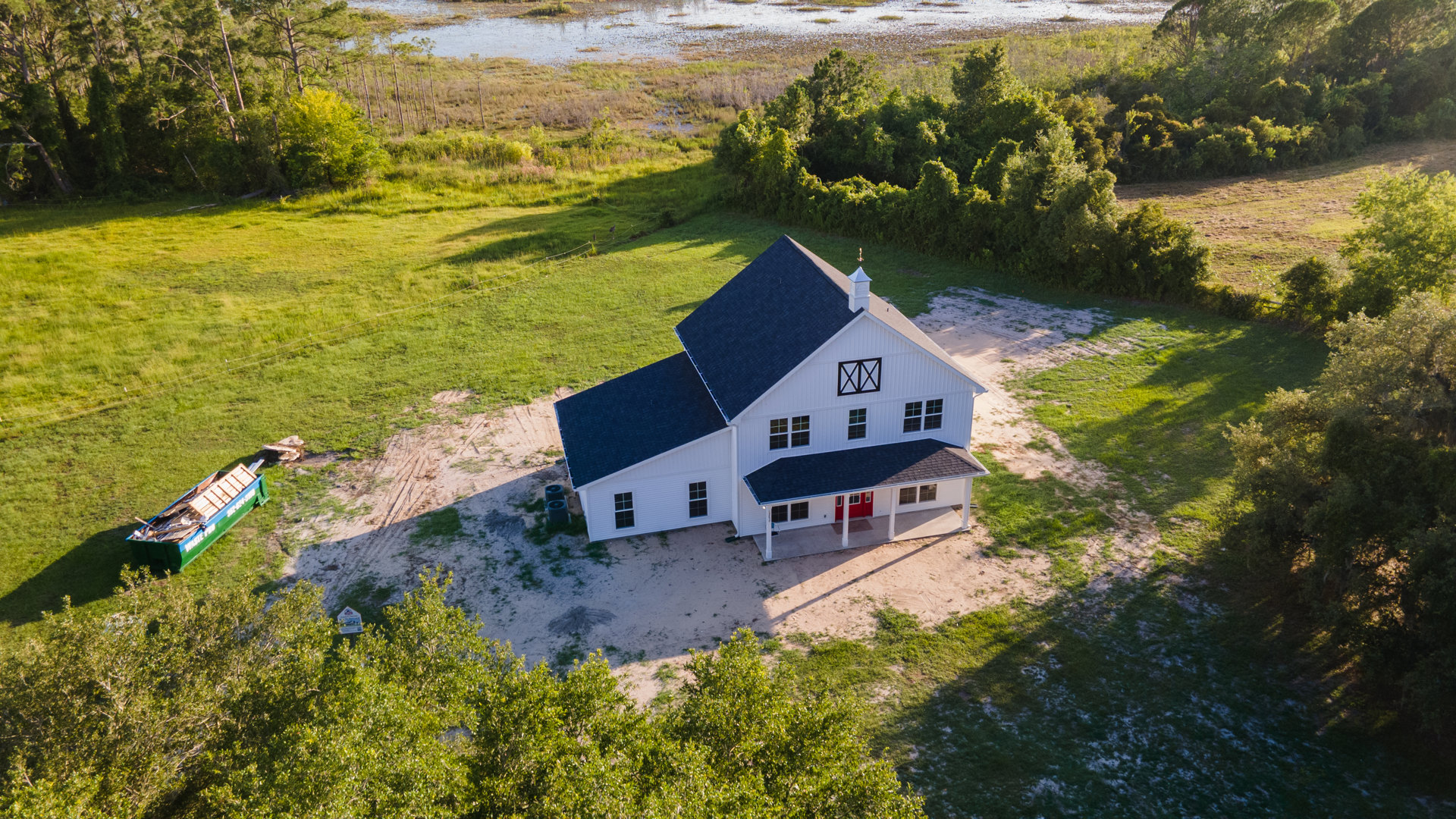 White house with blue roof, red door, and lawn, surrounded by trees and grass; green and blue dumpster filled with wood near the exterior.