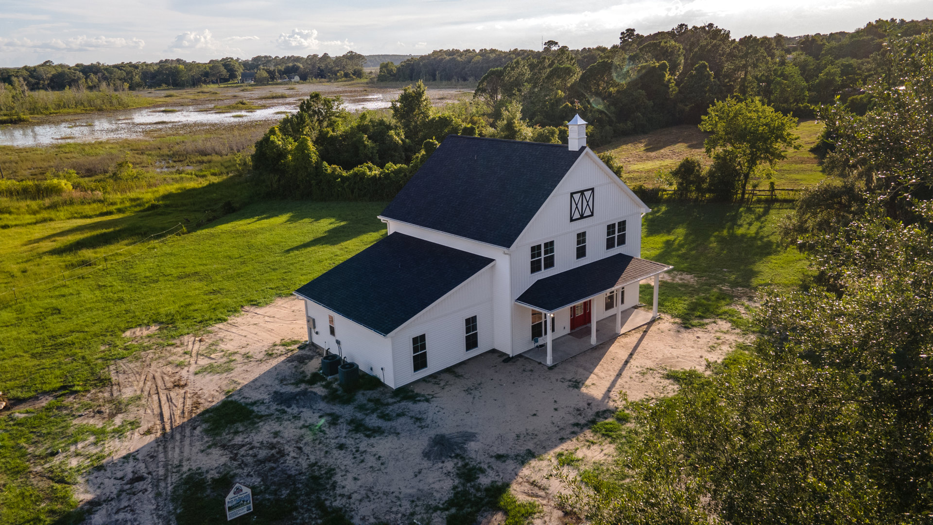 White siding house with red front door, expansive green lawn, and distant trees under partly cloudy sky