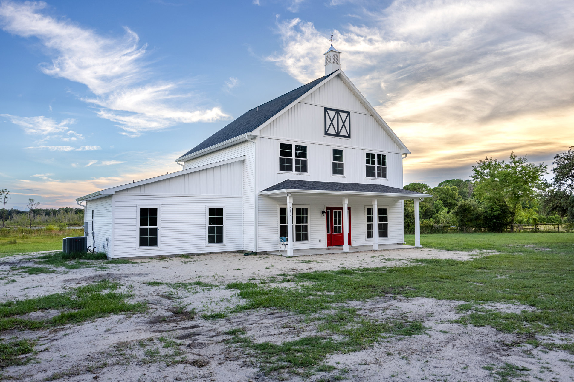 White farmhouse with red front door, black roof, white-framed windows, grassy lawn, and mature trees under a cloudy sky