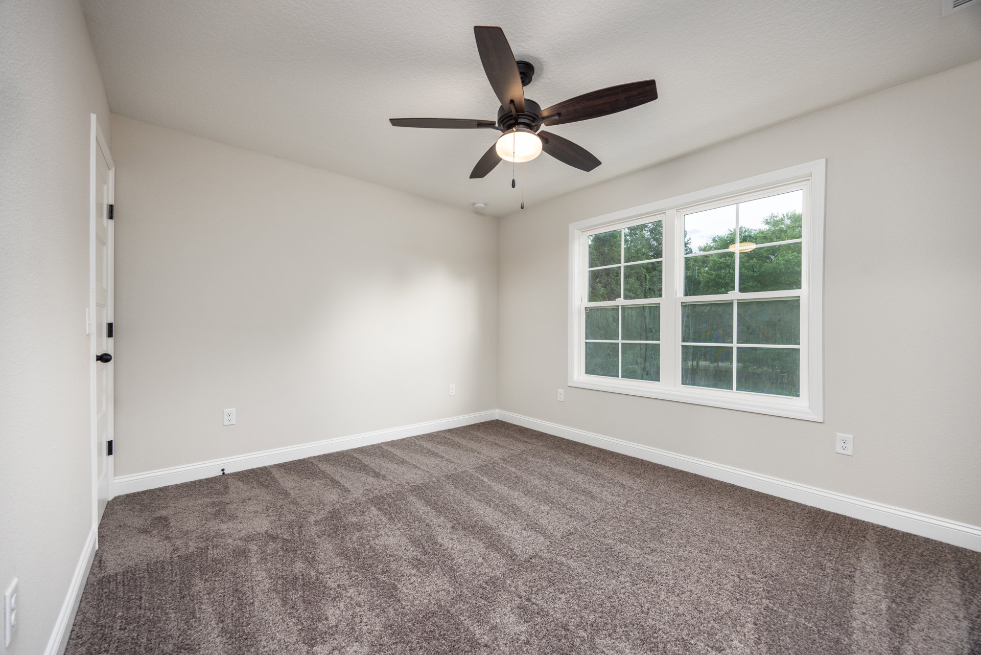 Ceiling fan with integrated light fixture in a carpeted room featuring white walls and a large window framed in white, overlooking trees outside