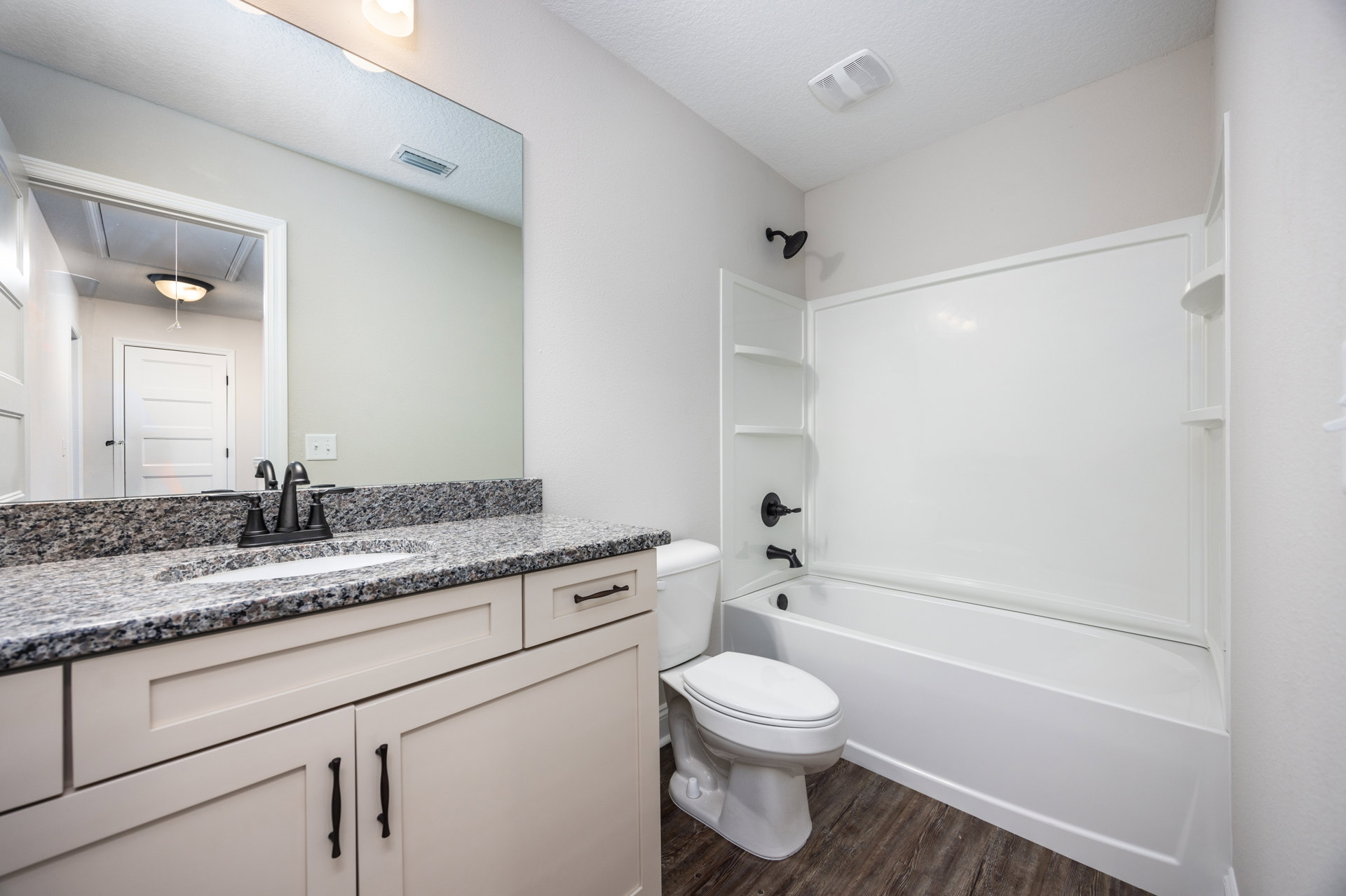White bathtub and toilet against light tile walls, black hardware on white door, chrome faucet, white ceiling vent, neutral cabinetry and countertop
