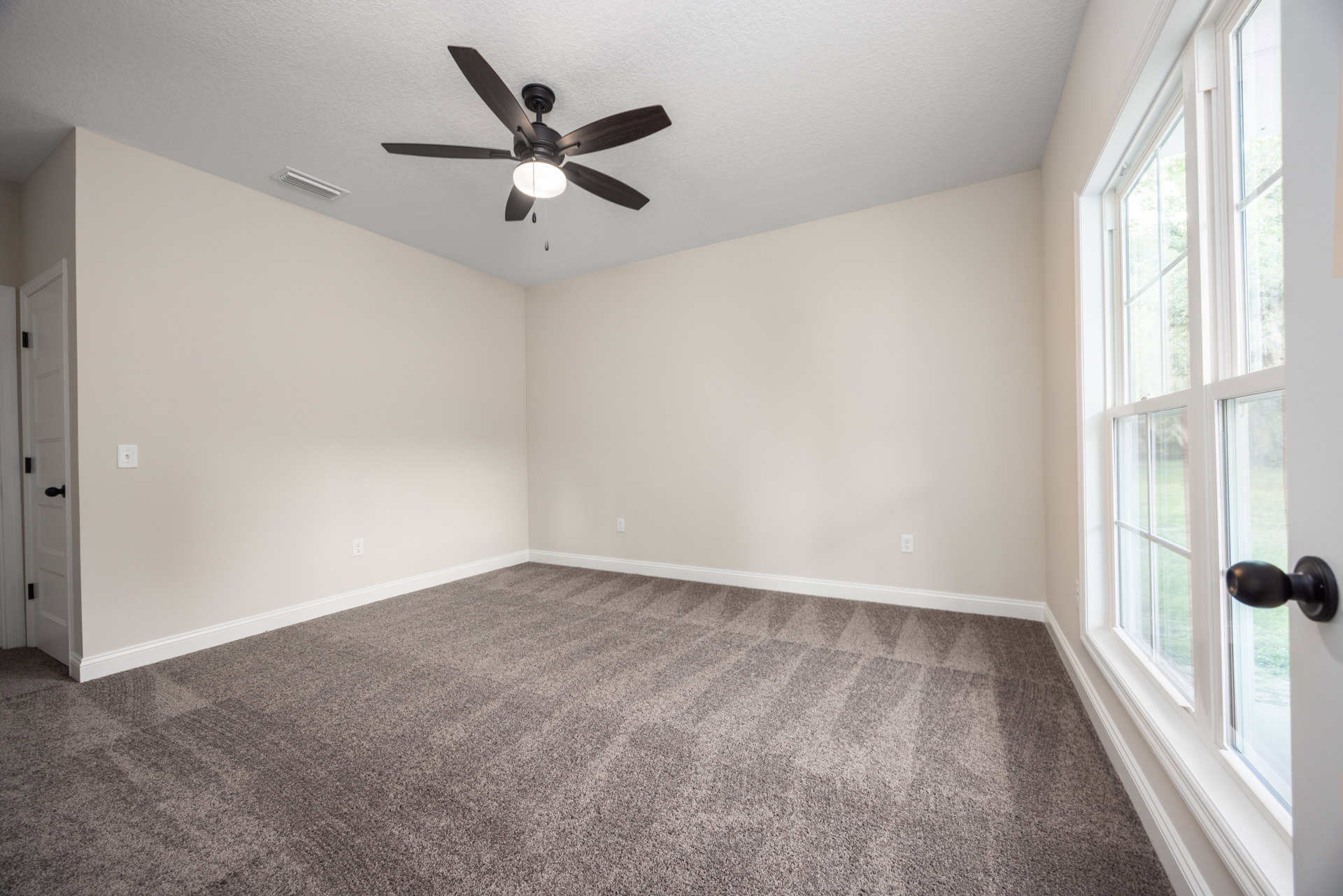Neutral-toned room with carpet flooring, white walls, ceiling fan with integrated light, large window, and paneled door with brushed metal knob