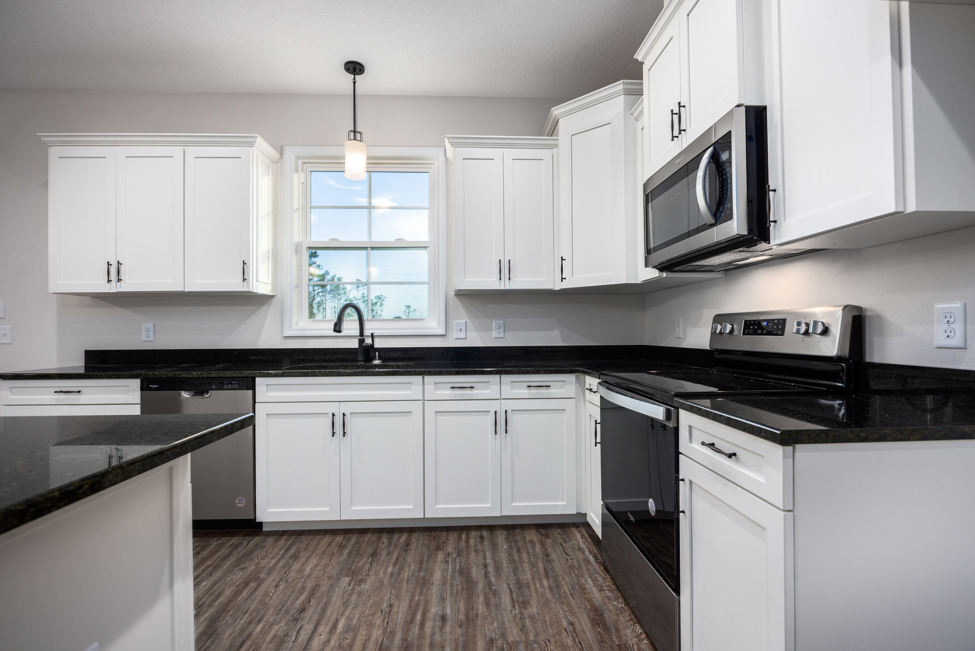 White kitchen cabinets with black countertops, stainless microwave set in cabinetry, wood flooring, black faucet beneath a white-trimmed window, electrical outlet visible near
