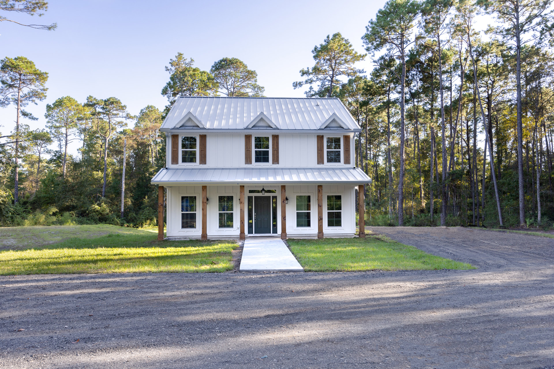 White house with black-framed windows and door, concrete driveway and walkway, wooden porch posts, mature trees and grass in background