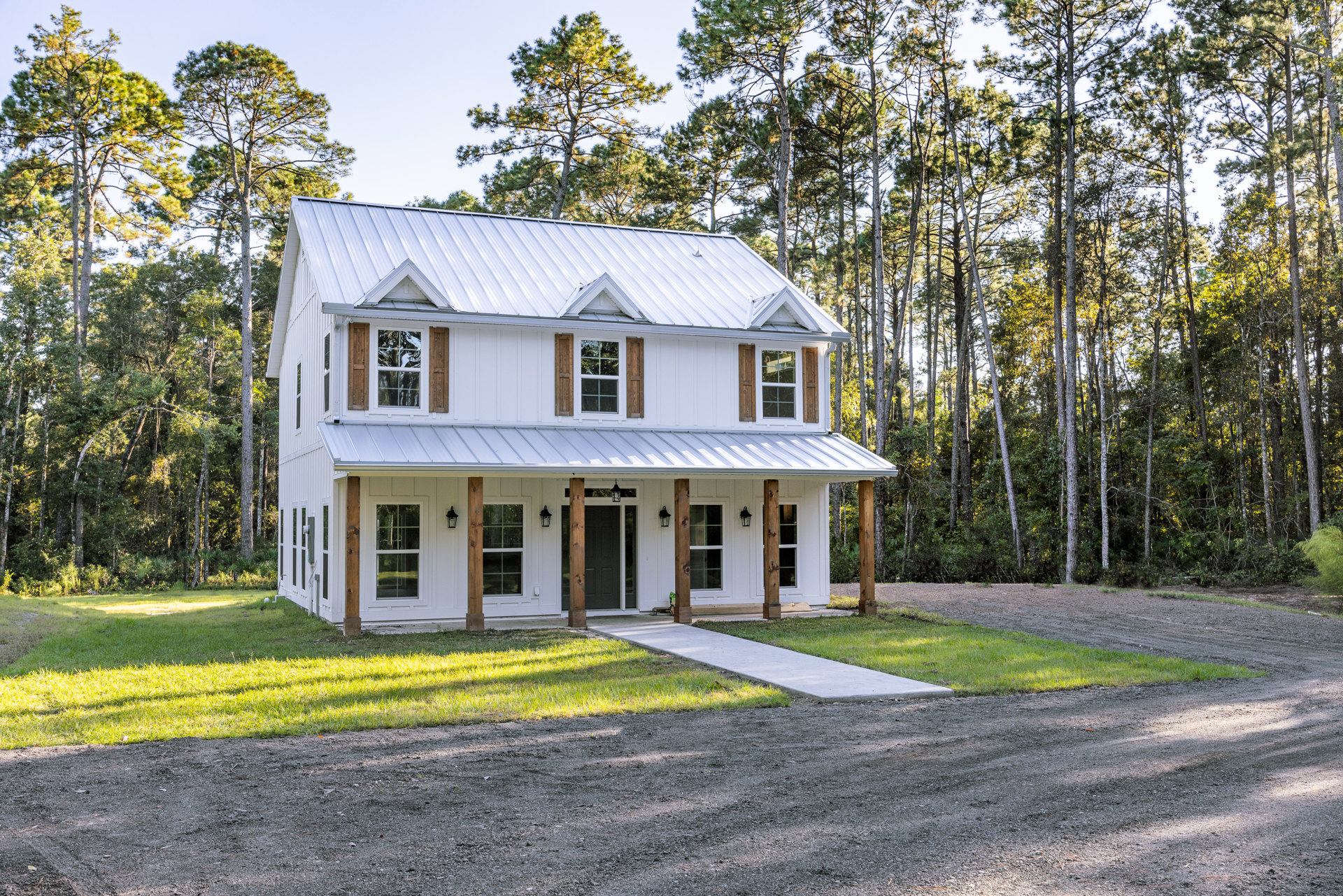 White house with wood pillars on the porch, concrete driveway and walkway bordered by grass, mature trees in the background, windows with shutters on the side.