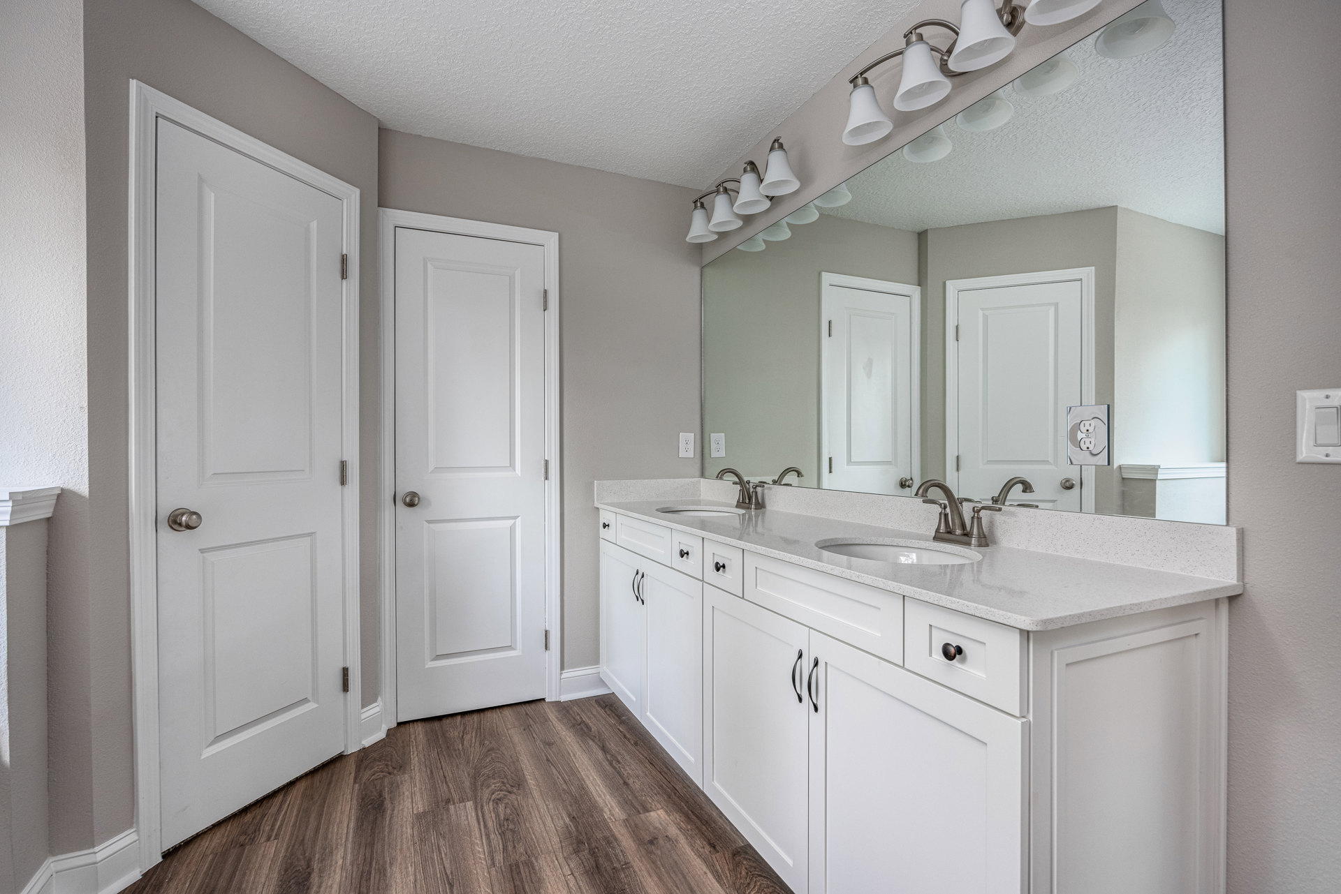Bathroom with white shaker cabinets, white paneled doors with silver hardware, wood flooring, wall-mounted light fixture above a rectangular mirror, and neutral tile accents.