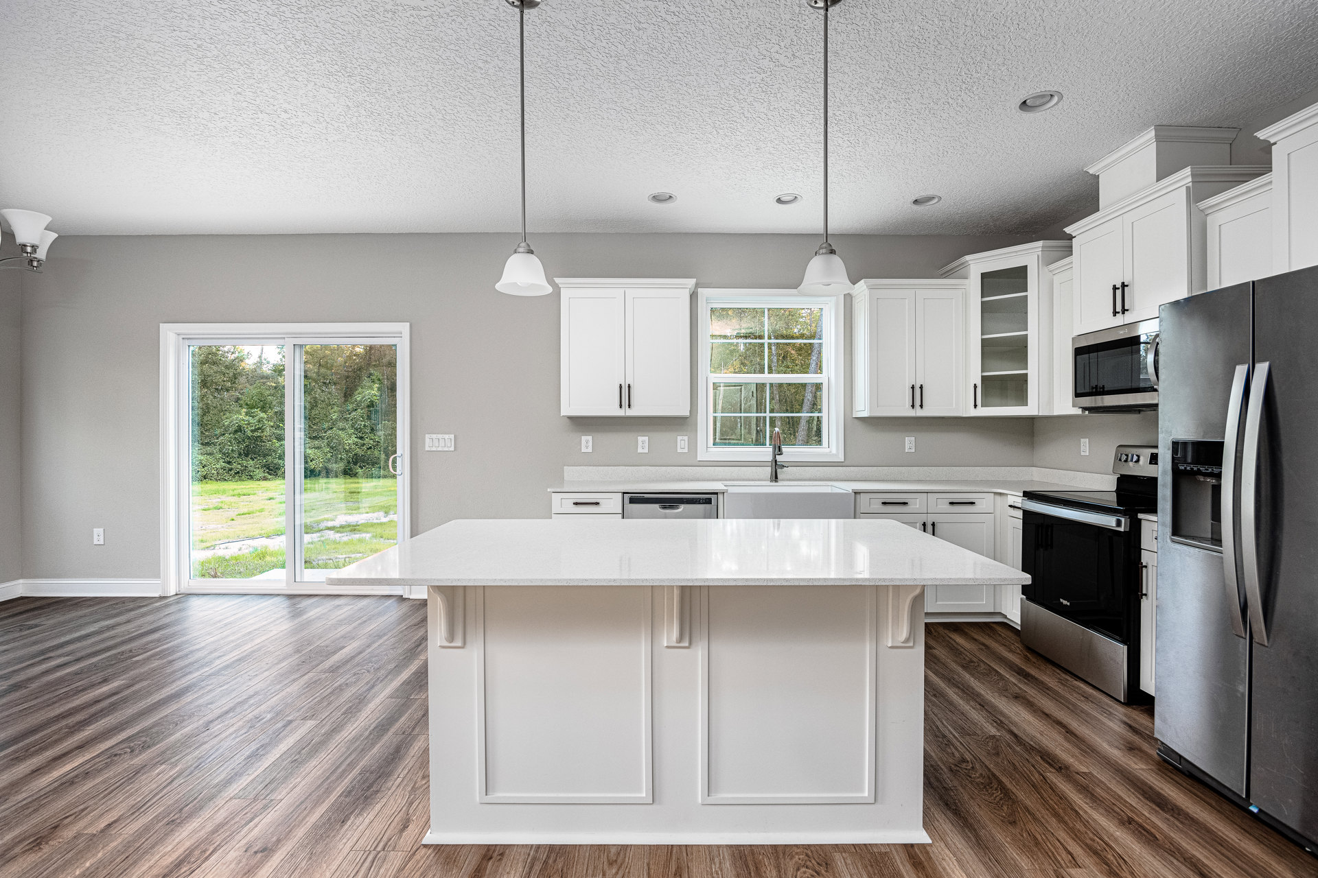 White kitchen with shaker cabinets, large central island, quartz countertops, stainless steel appliances, window overlooking green lawn and trees.