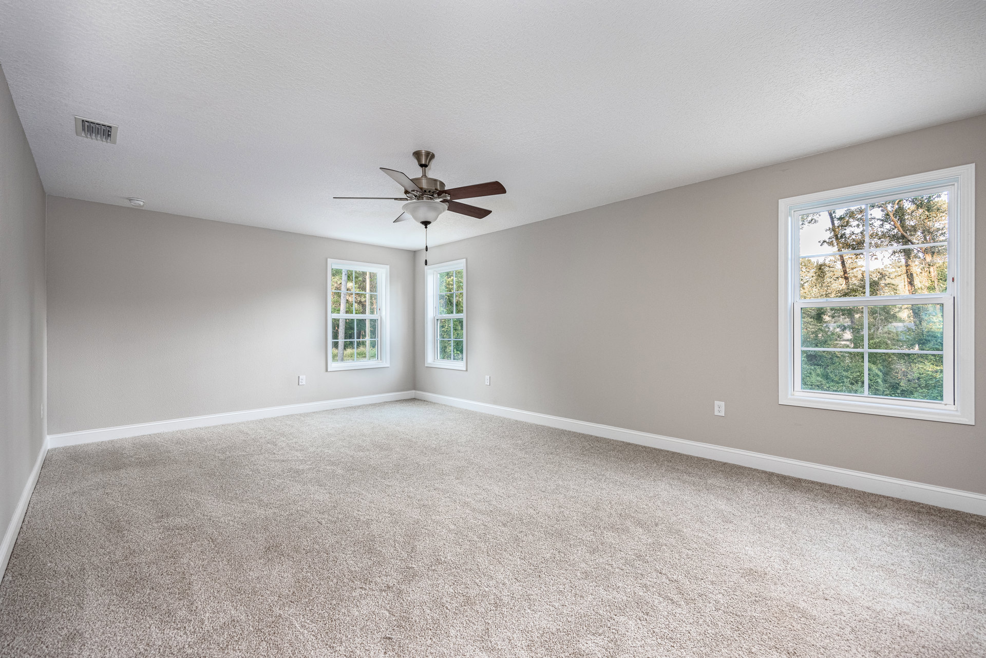 White carpeted room with white walls, ceiling fan with light fixture, large window overlooking green trees, simple molding along ceiling and floor.