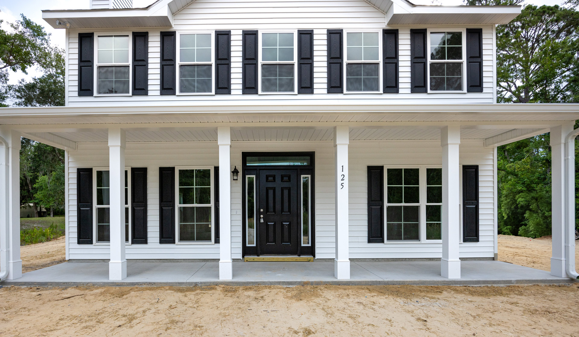 White siding house with black shutters, black front door with glass panels, white-framed windows, covered porch, and mature trees in front yard