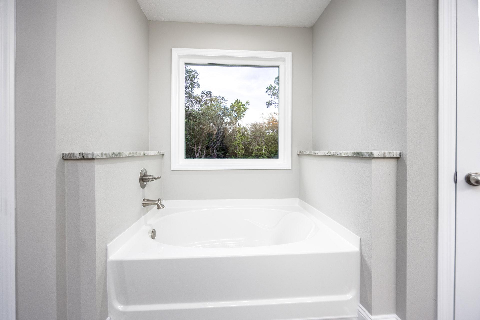 Freestanding white bathtub beneath large window with tree views, surrounded by light tile flooring and white walls, chrome faucet and door knob visible